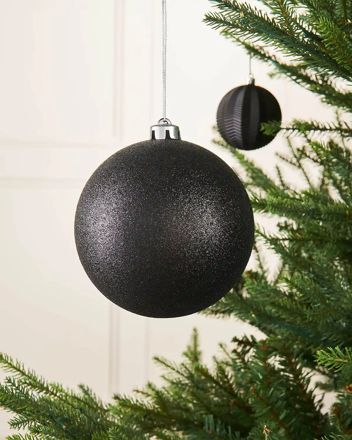 Black glitter oversized 15cm bauble on a Christmas tree branch with a smaller black bauble in background