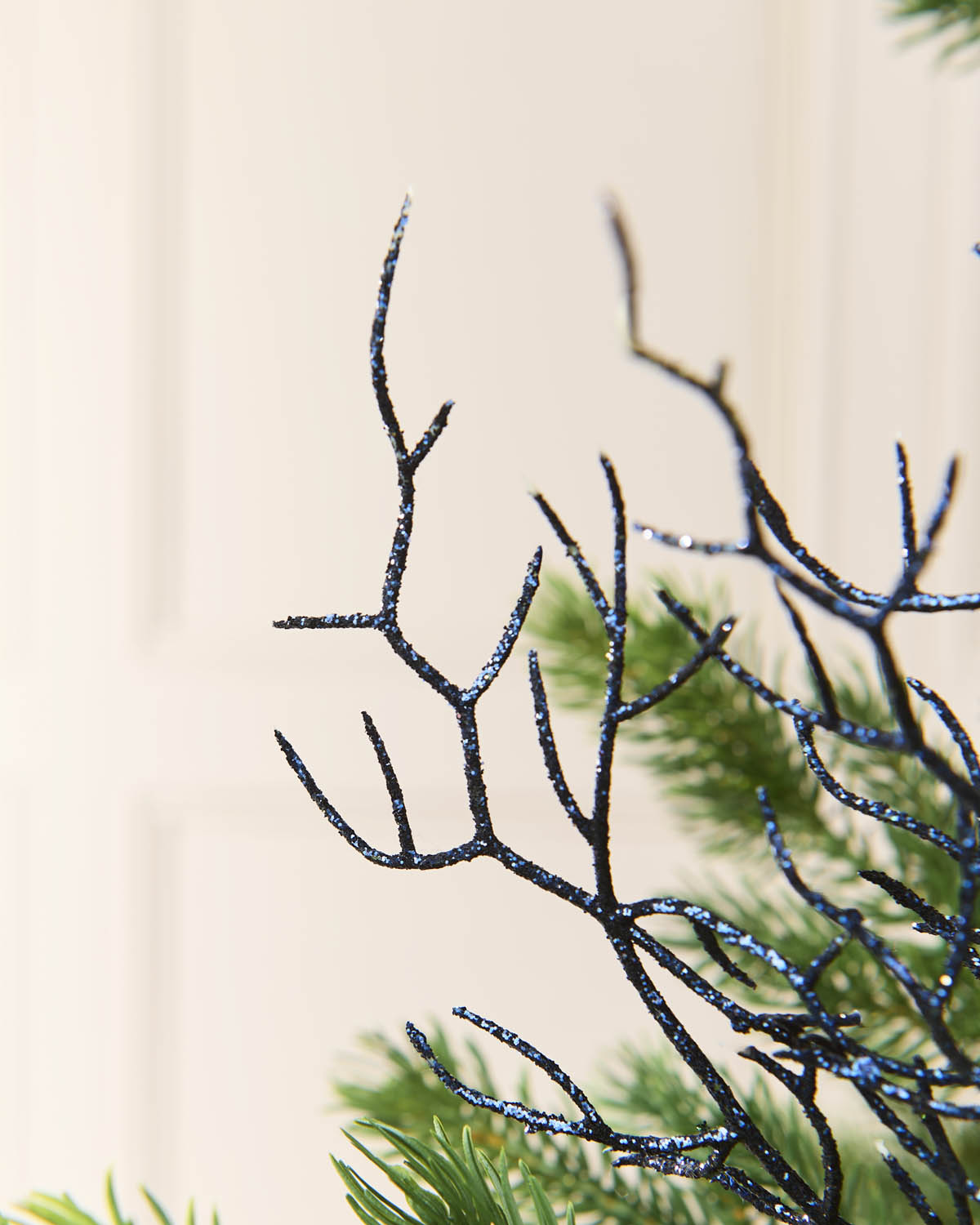 Black glittery coral-like decorative decor with blue glitter accents, shown against a soft cream background with green pine needles visible at the bottom.