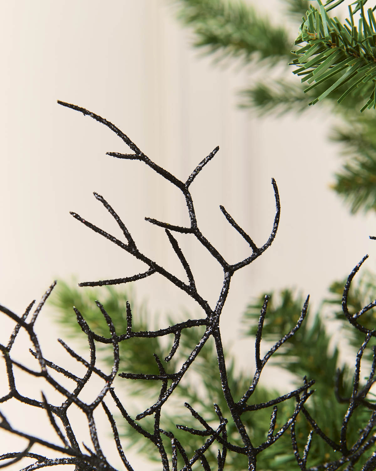 Black glittery coral-shaped holiday decoration with a sparkly finish, shown against a soft beige background with blurred green pine needles.