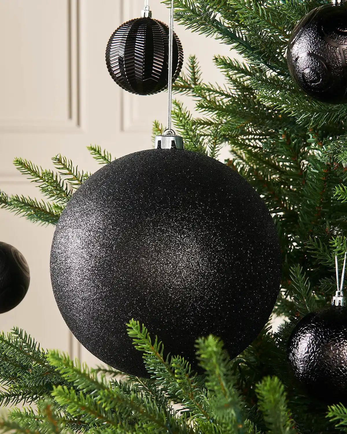 Black glittery oversized Christmas bauble on a Christmas tree branch with 4 smaller black baubles in background