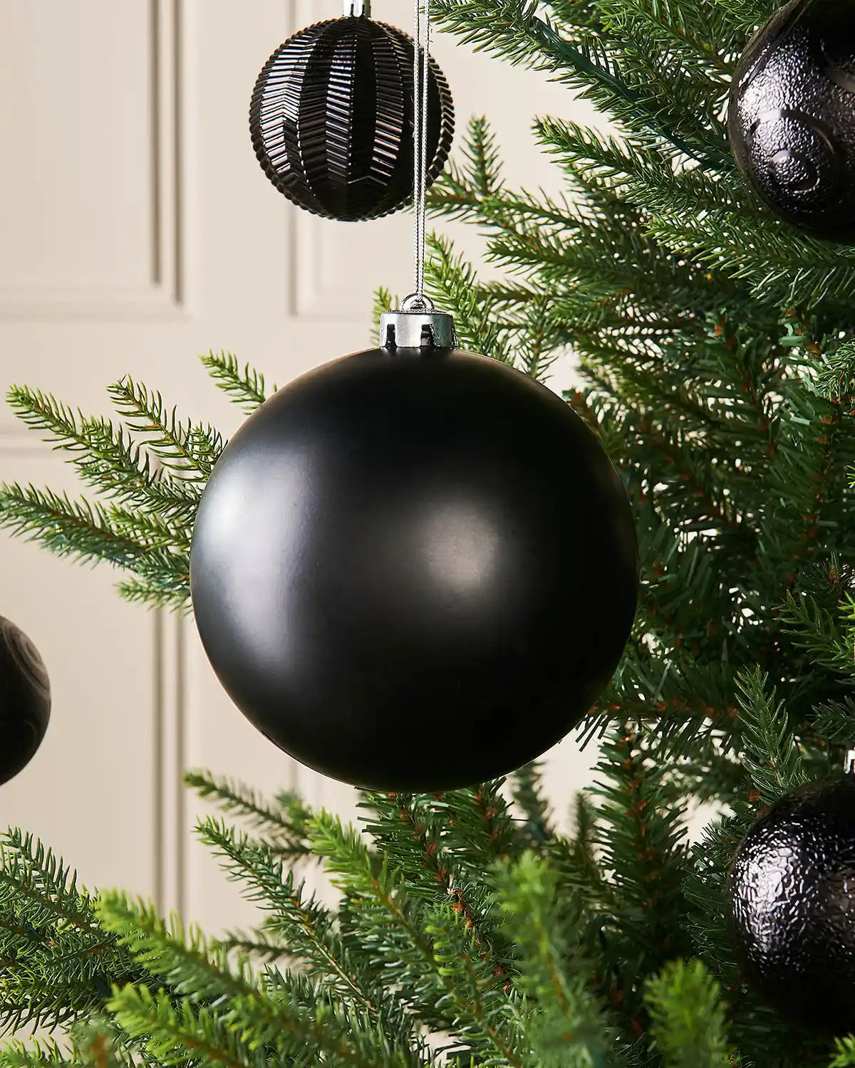 Black matt oversized 15cm bauble on a Christmas tree branch with 4 smaller black baubles in the background