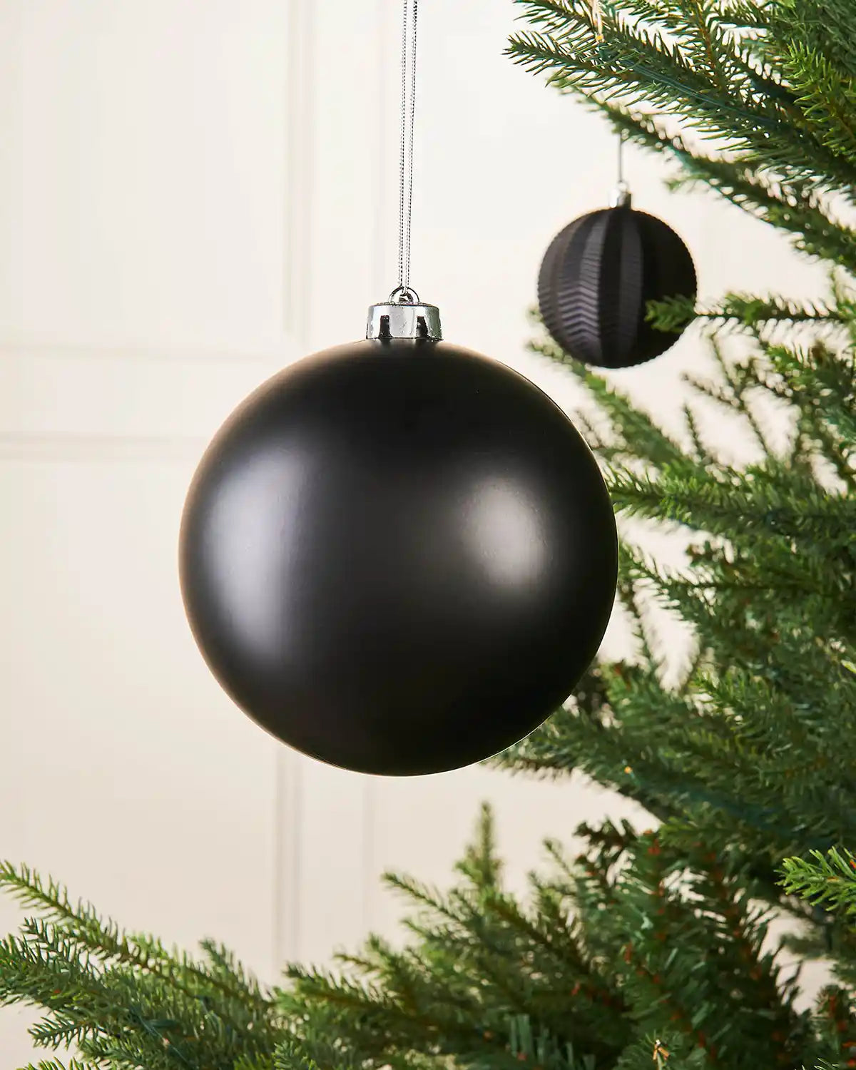 Black matt oversized 15cm bauble on a Christmas tree branch with a smaller black bauble in the background