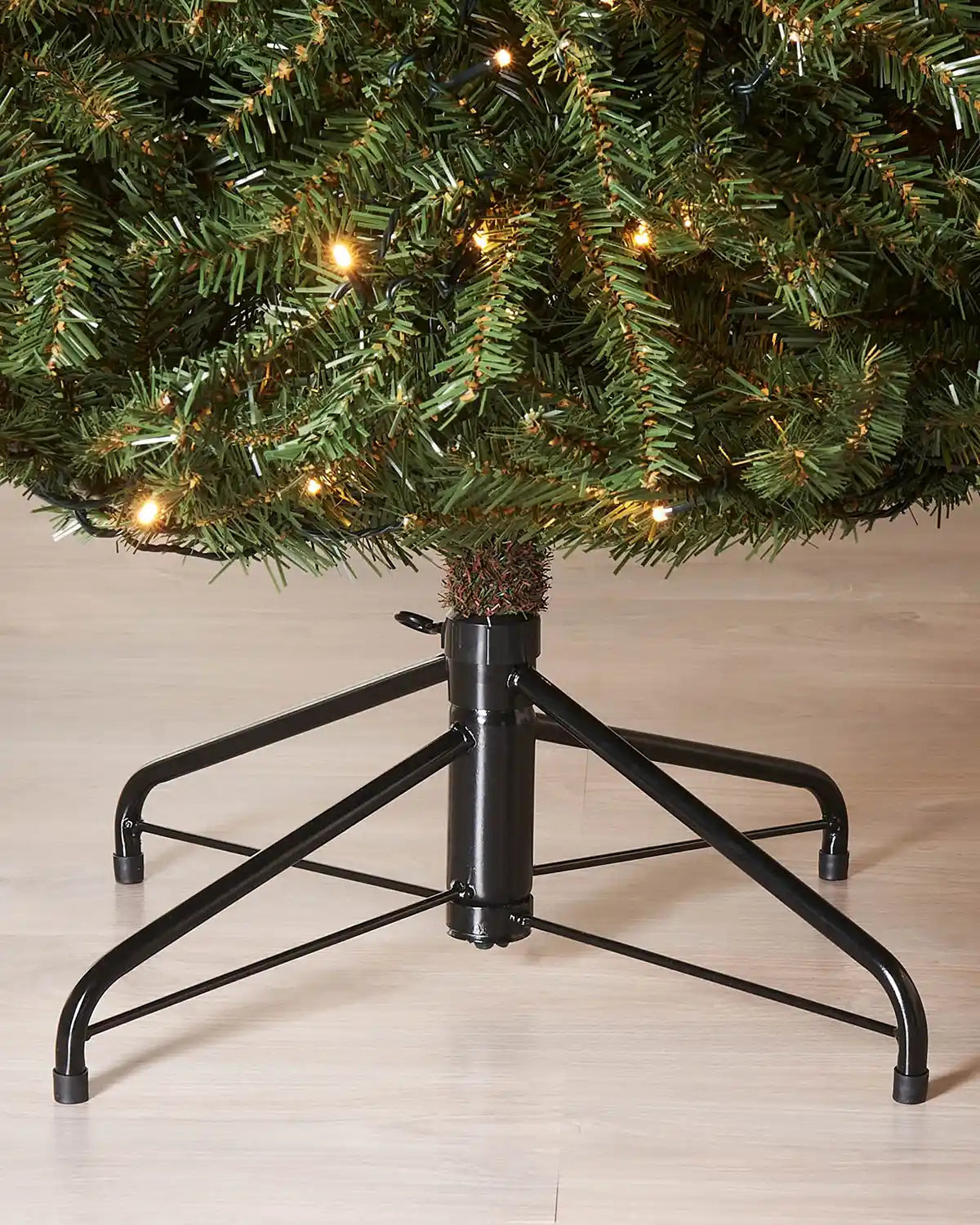 Black metal Christmas tree stand and base of 7tf with a Christmas tree on a light wooden floor.
