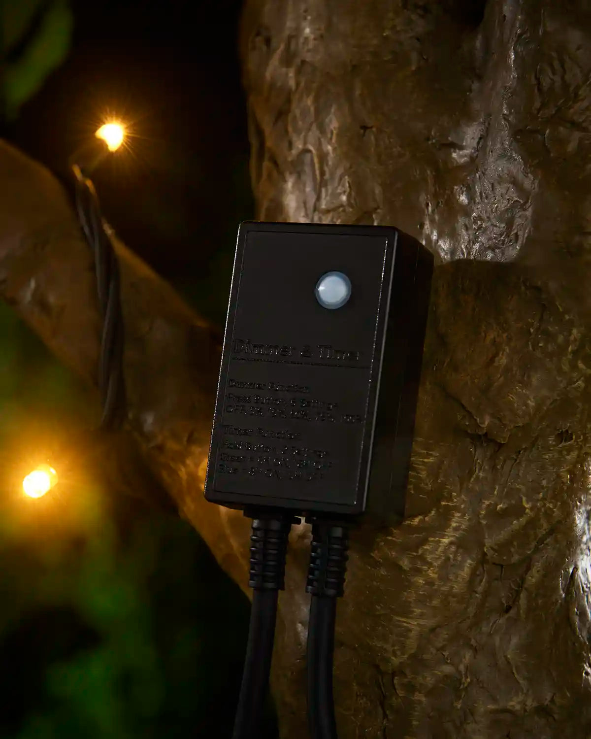 Black outdoor Christmas light dimmer/timer controller mounted against a tree, with warm white string lights glowing in the background.
