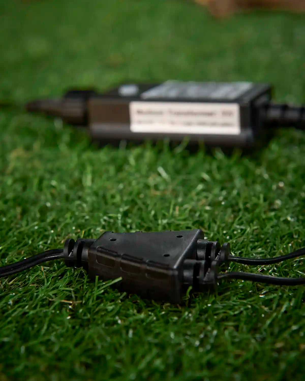 Black outdoor light splitter with cables on grass, with a transformer blurred in the background