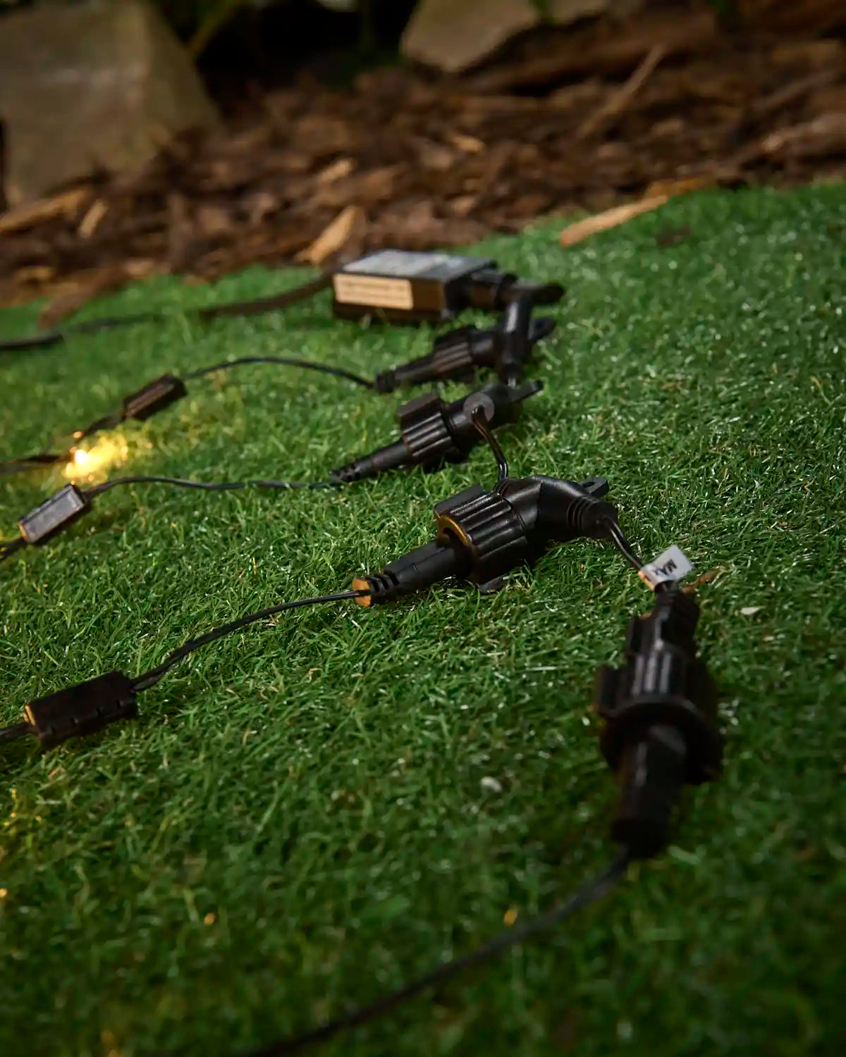 Black outdoor light splitters and connectors arranged on grass, with a transformer and warm white string lights in the background