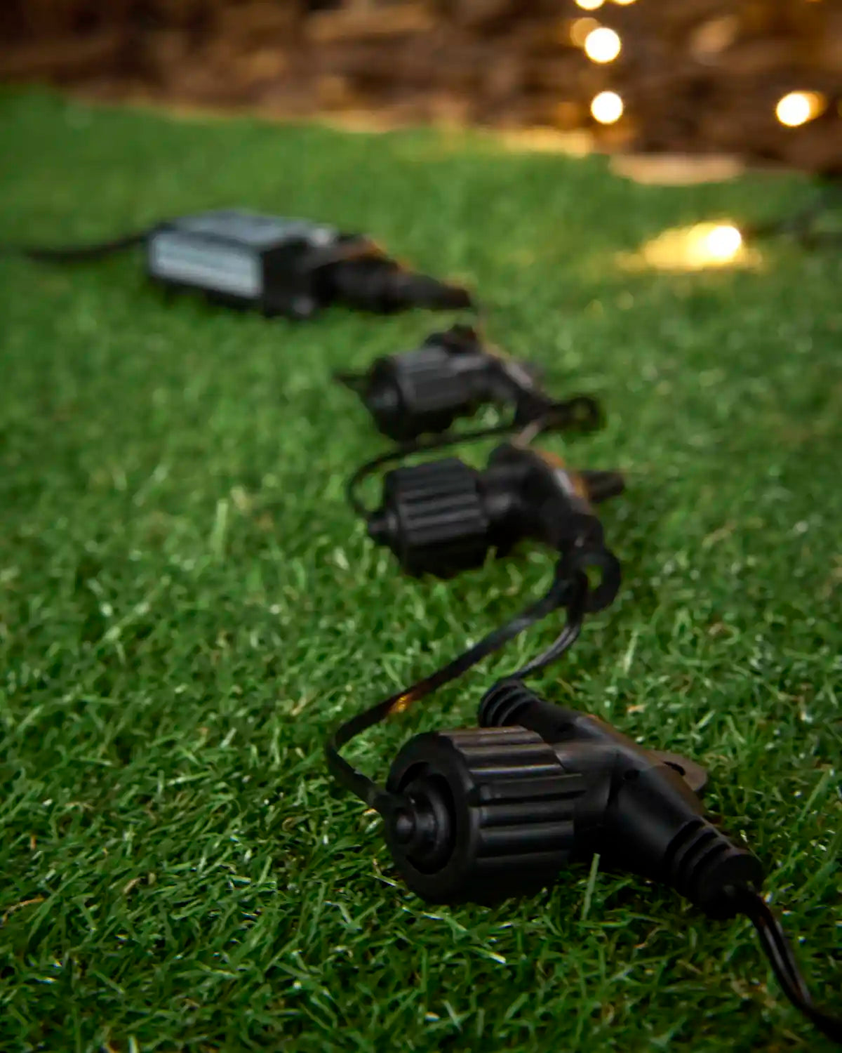 Black outdoor light splitters connected in a line on grass, with warm white string lights glowing in the background