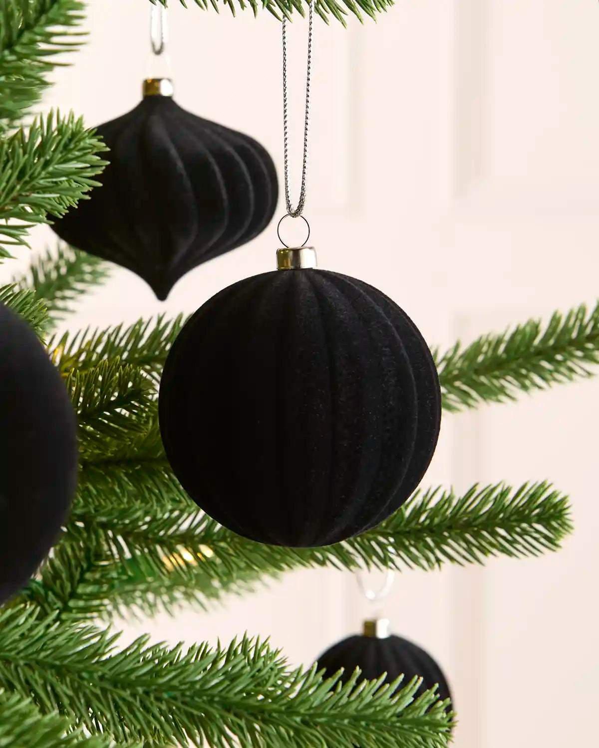 Black ribbed velvet Christmas tree ornaments hanging from green pine branches against a soft cream background.