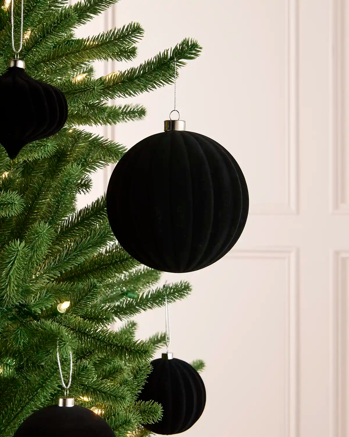 Black ribbed velvet Christmas tree ornaments hanging on a green Christmas tree against a light cream paneled wall.