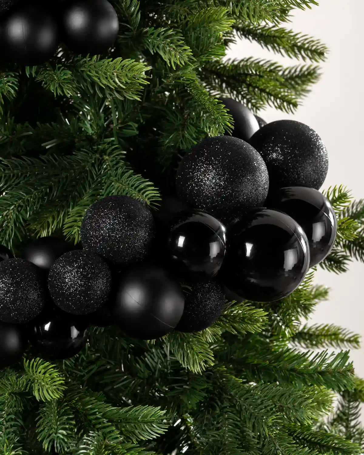 Black spherical Christmas tree ornaments, including some matte black and some glittery black, draped over a green artificial pine tree against a light neutral background.