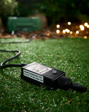 Black 6W transformer for outdoor LED string lights on grass, with warm white lights glowing in the background.