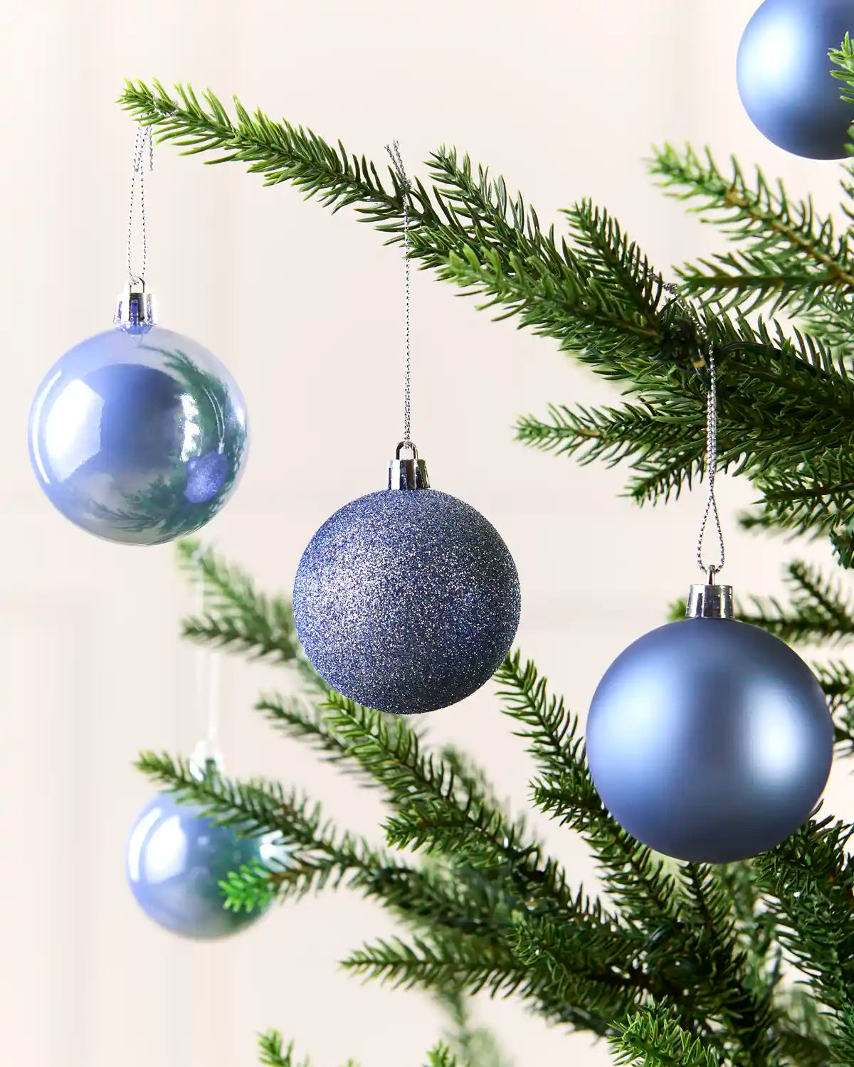 Blue Christmas tree ornaments hanging on green pine branches against a soft cream background.