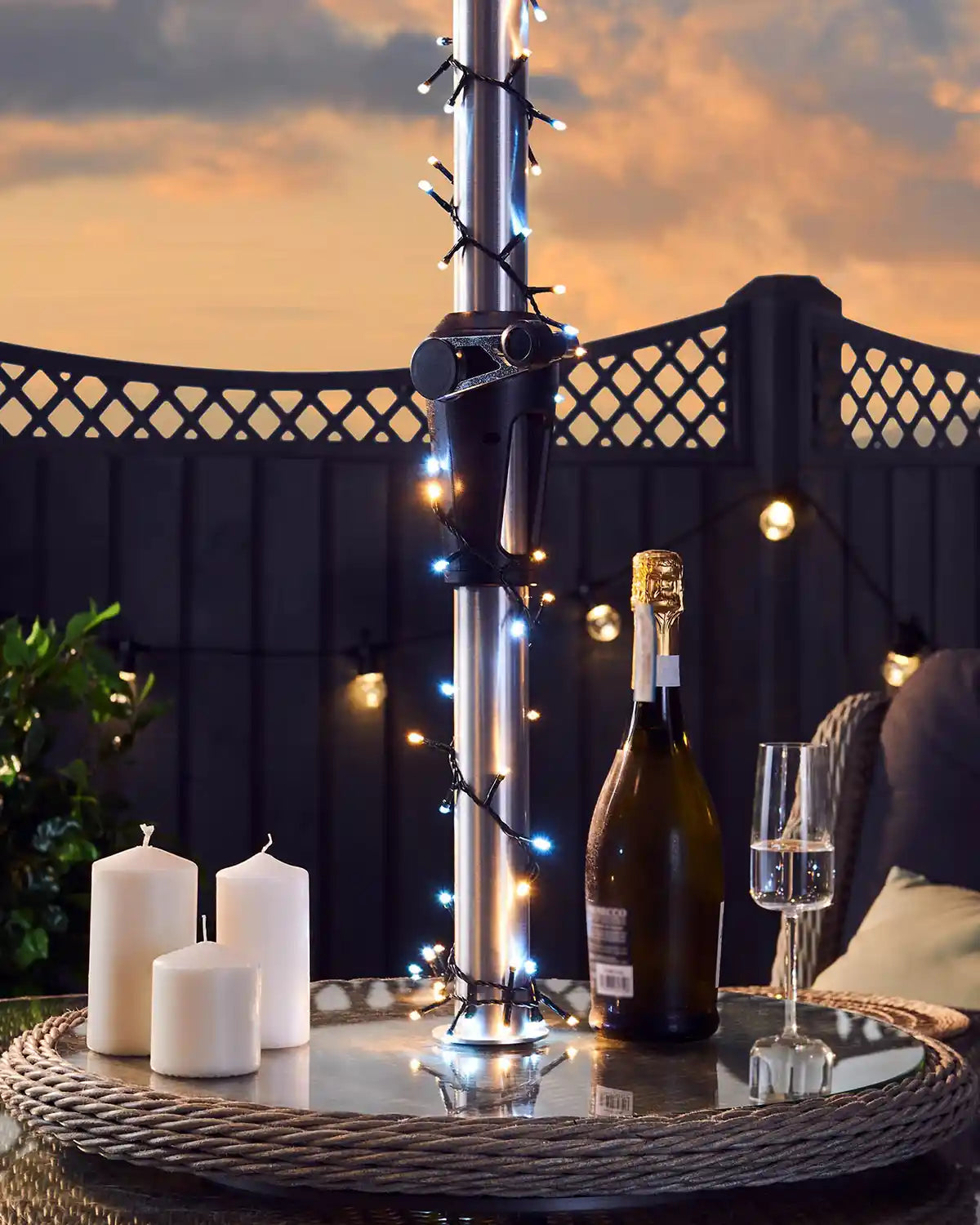 Bottle of champagne with a glass on a table and Warm white and ice white fairy lights on an umbrella outdoors at sunset.