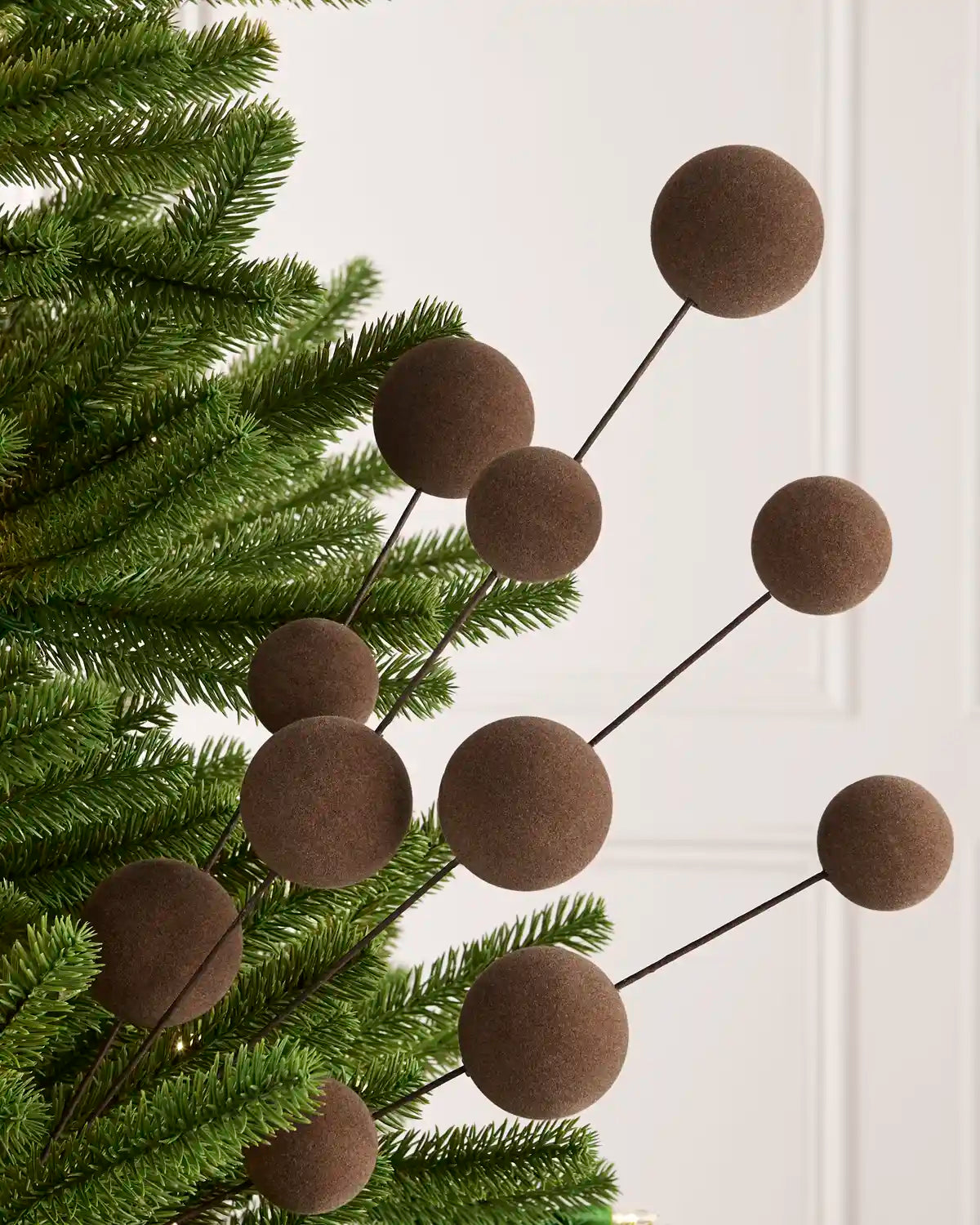 Brown felt ball branch garland with dark metal stems, shown draped over green Christmas tree branches against a white paneled wall.