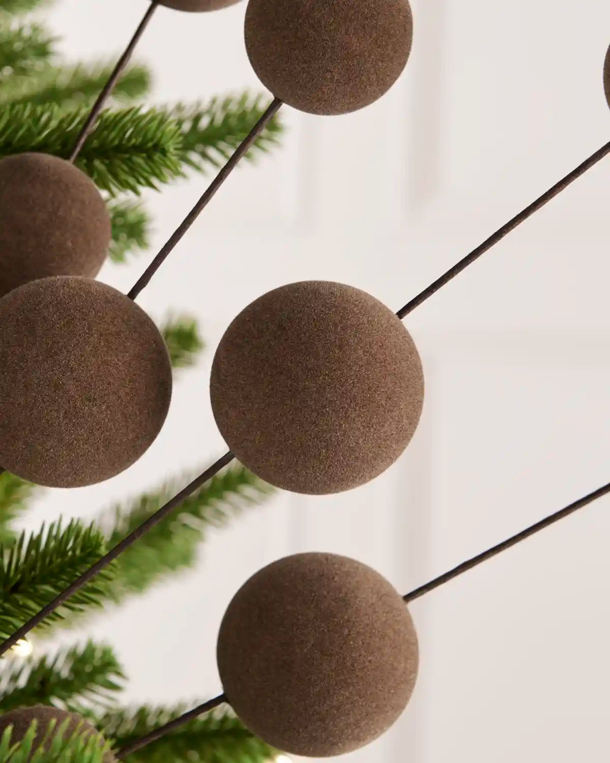 Brown felt ball garland hanging on thin black wires, shown with green pine branches against a soft white background.