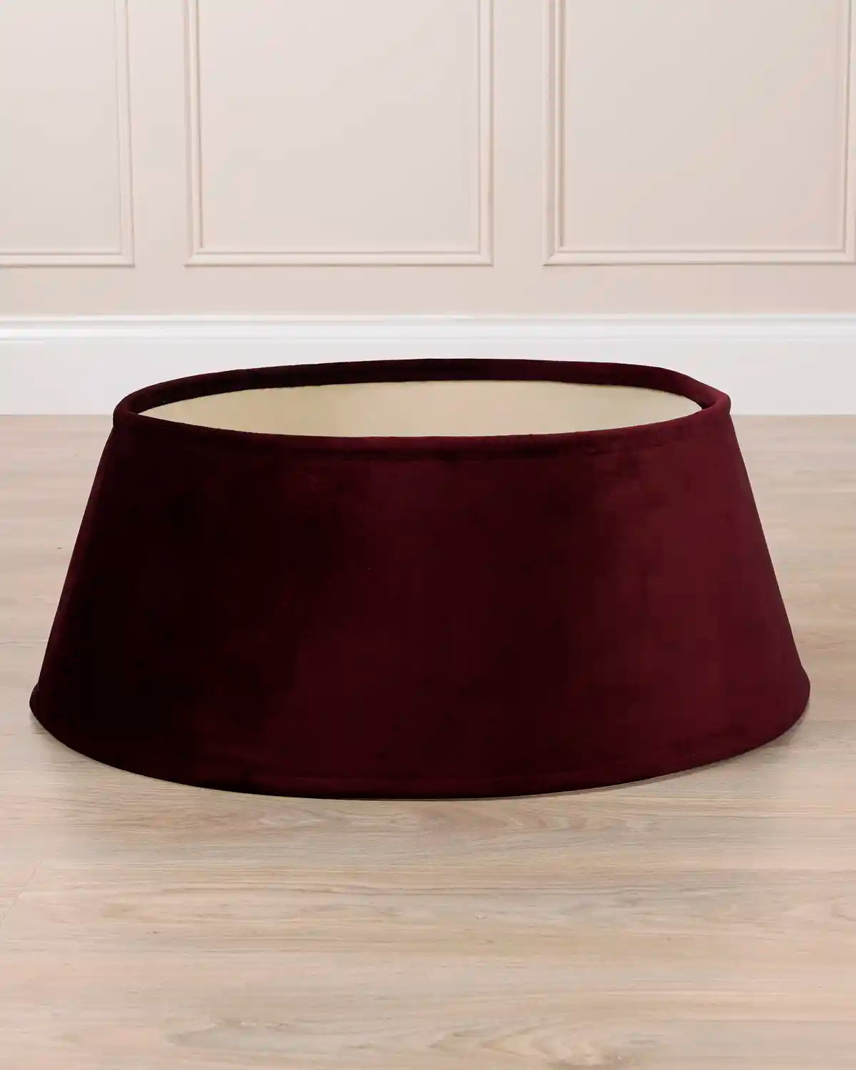 Burgundy coloured velvet tree skirt with a wide, flared dome shape, shown on a light wood floor against a white panelled wall.

