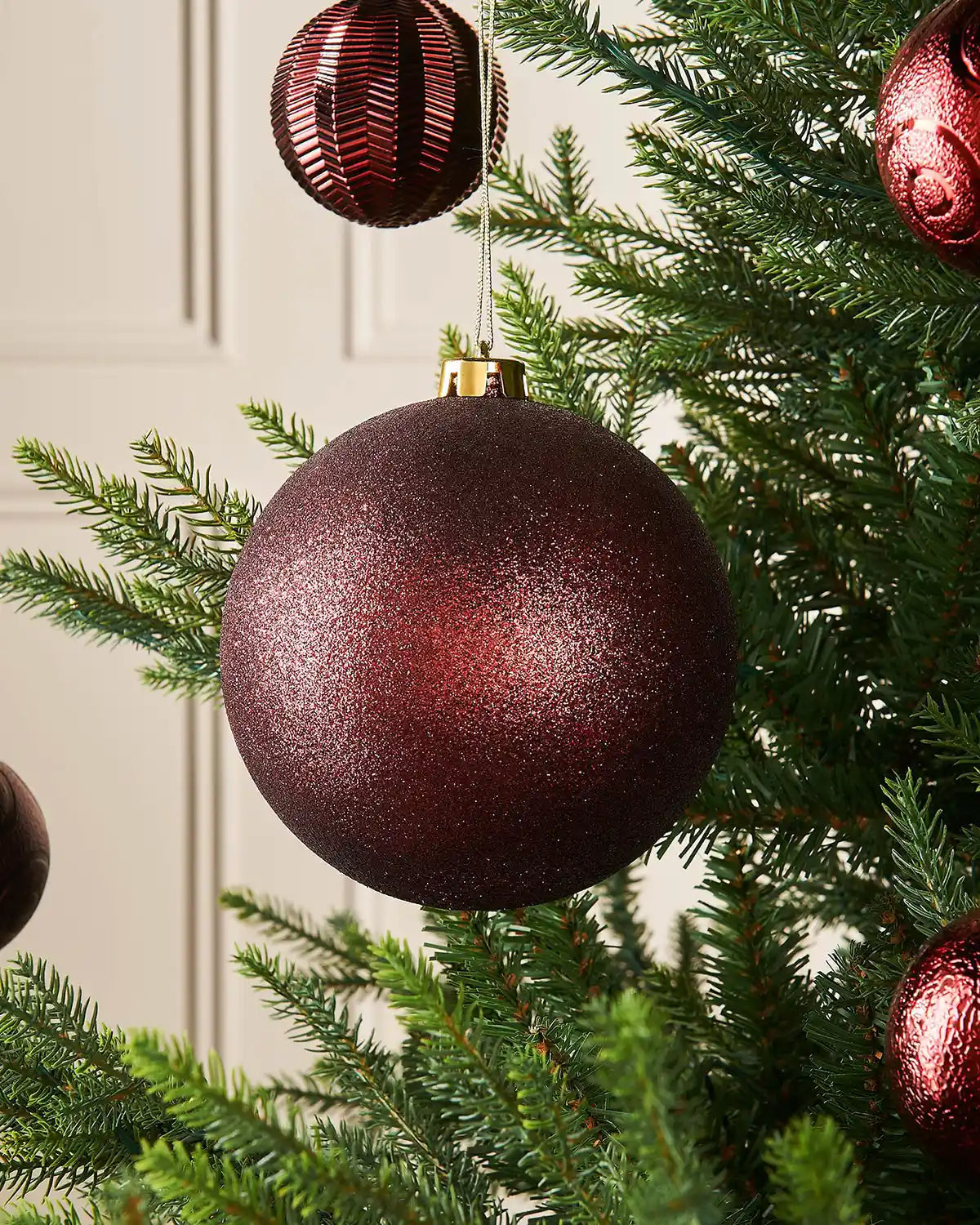 Burgundy glitter oversized 15cm bauble on a Christmas tree branch with 4 smaller burgundy baubles in the background