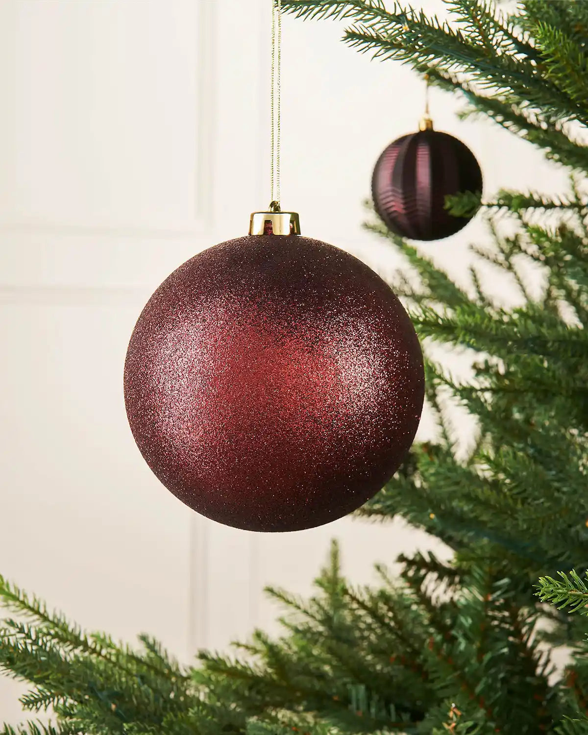 Burgundy glitter oversized 15cm bauble on a Christmas tree branch with a smaller burgundy bauble in the background