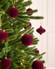 Burgundy velvet Christmas tree ornaments hanging on a green artificial Christmas tree with warm white string lights, set against a soft cream panneled wall.