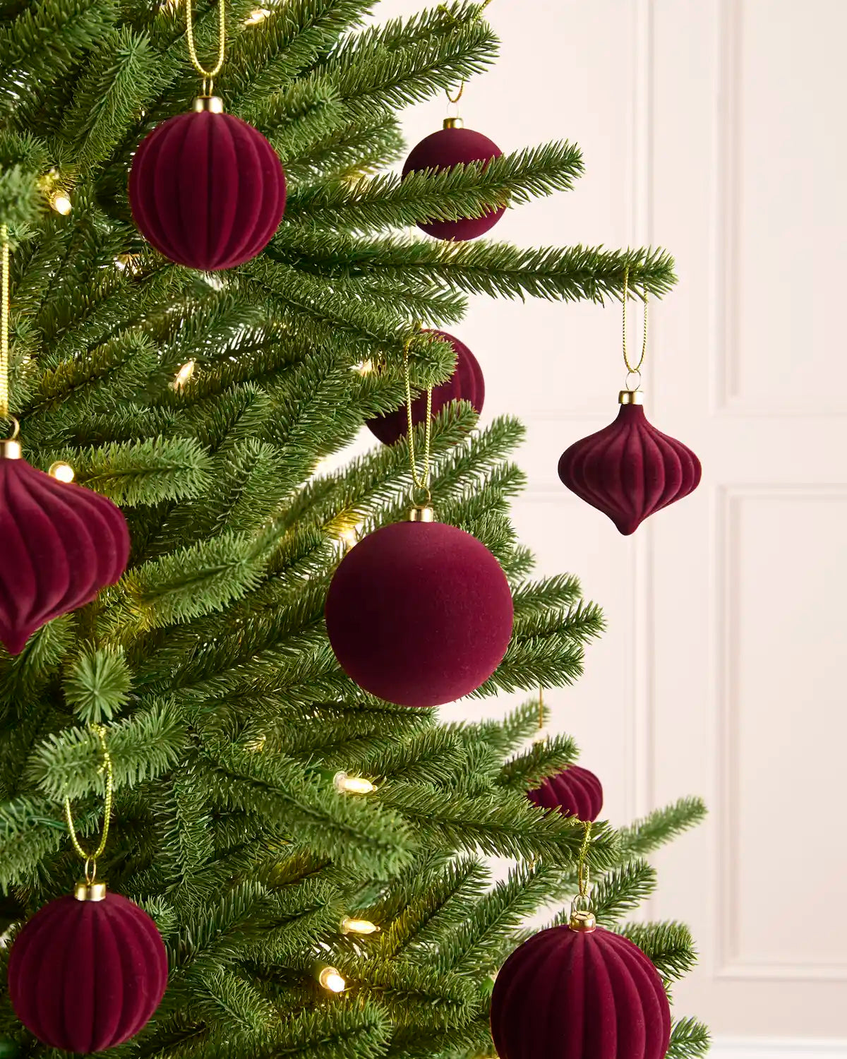 Burgundy velvet Christmas tree ornaments hanging on a green artificial Christmas tree with warm white string lights, set against a soft cream panneled wall.