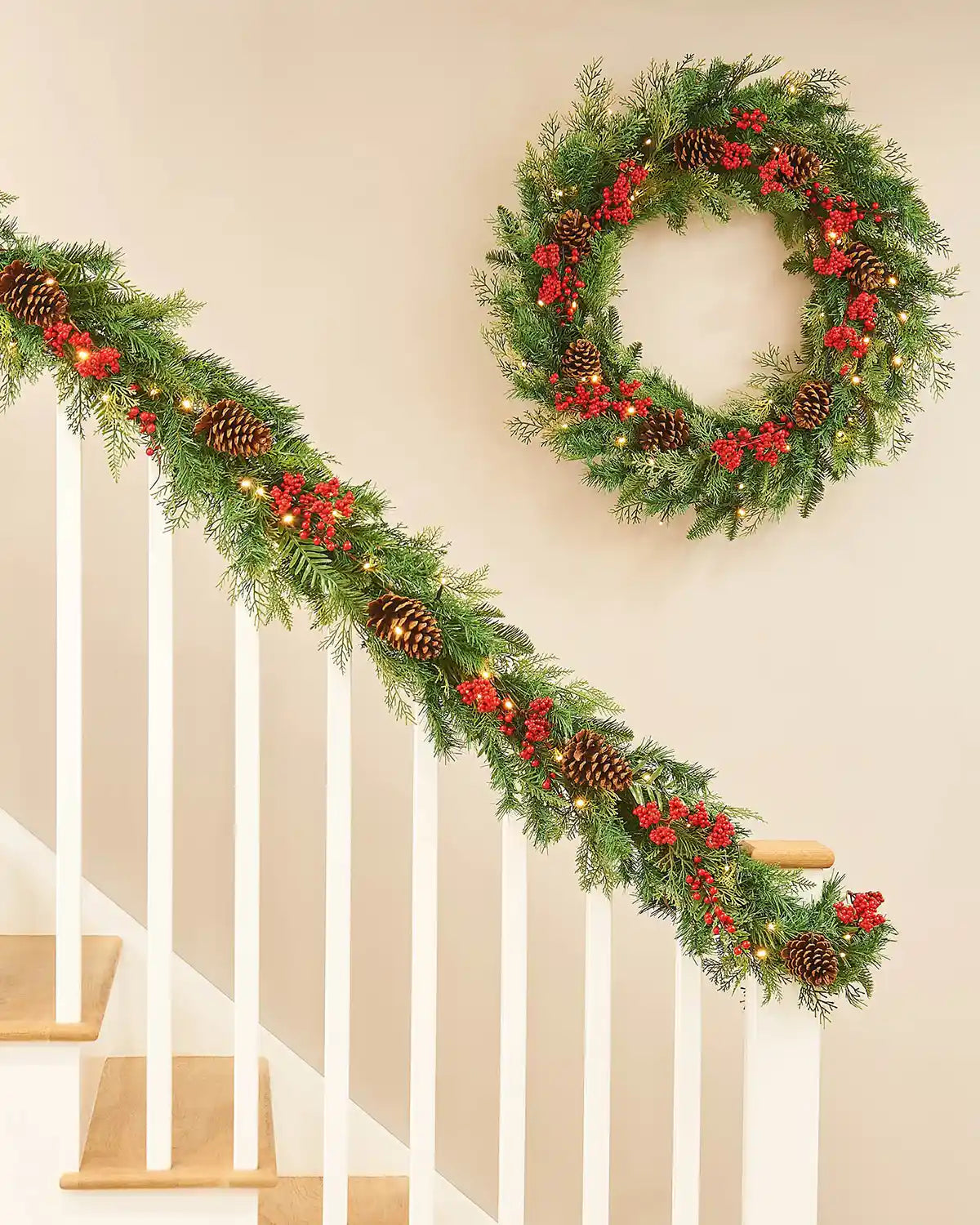 Christmas garland and matching wreath with greenery, red berries, and pinecones on a wooden staircase banister and a cream wall
