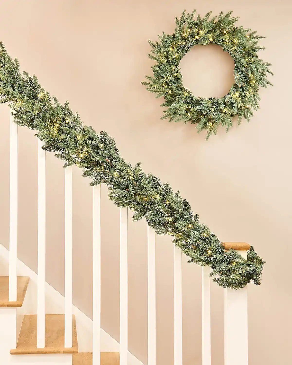 Christmas garland with a matching wreath with warm white lights on a staircase banister against a cream wall