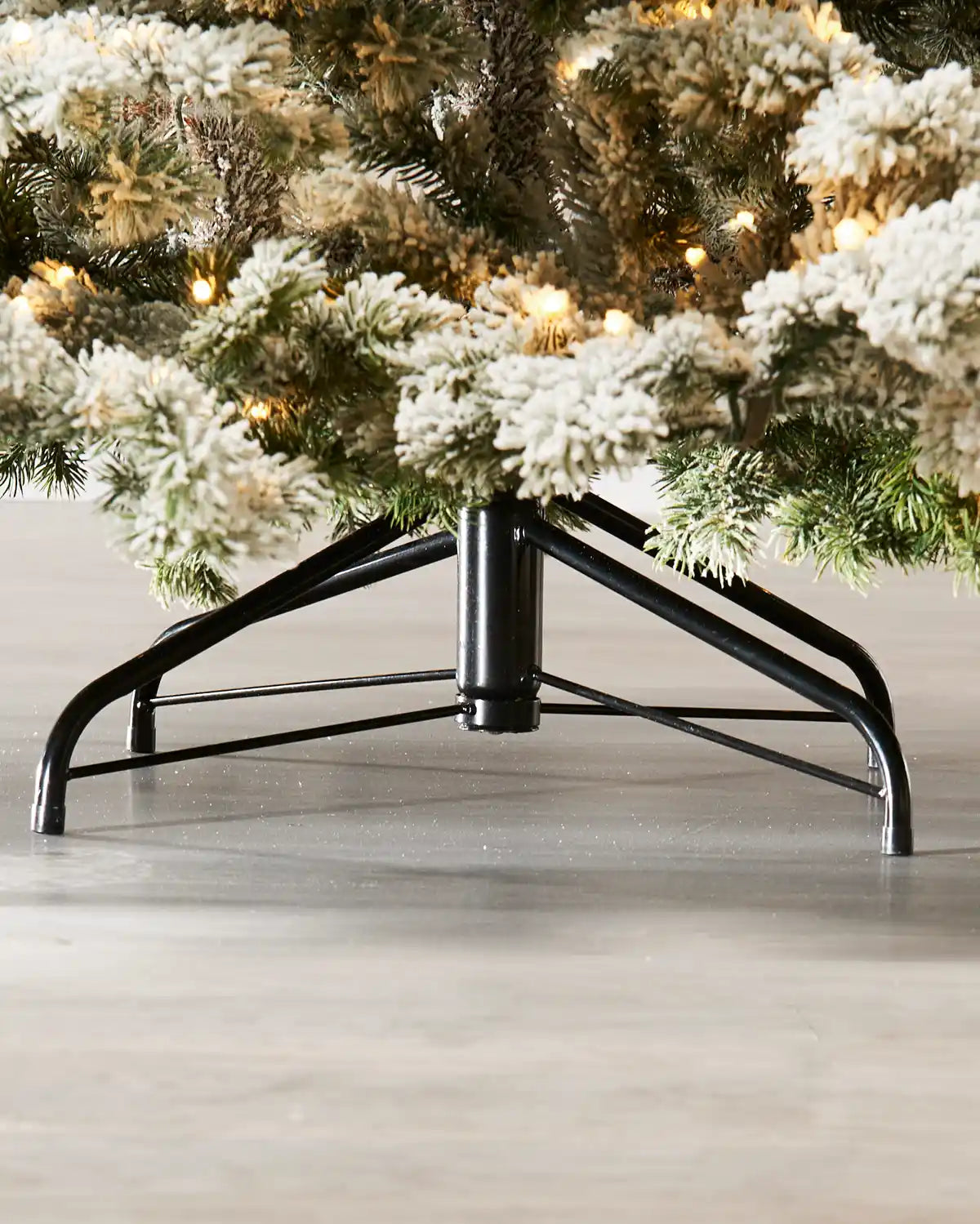 Christmas tree’s lower branches and sturdy black metal stand, featuring snow-flocked green needles and warm white lights above a light wood floor.
