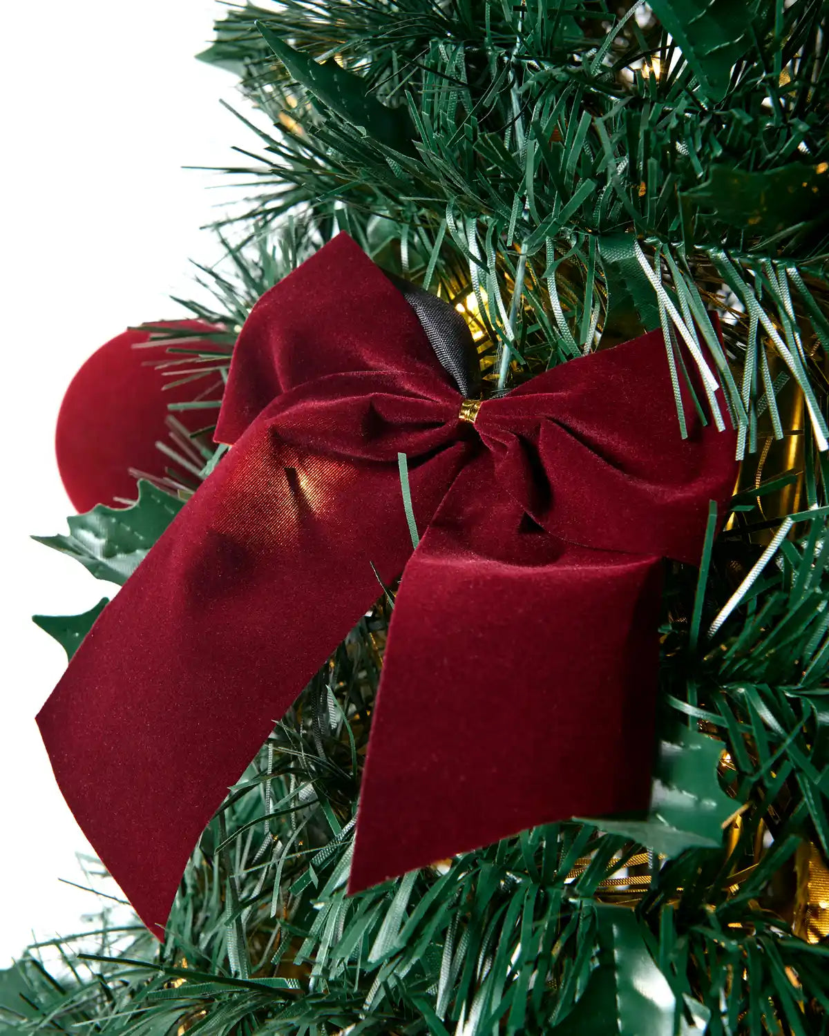 A dark green Christmas tree stand with a triangular base, shown supporting a decorated tree with a red bow and warm string lights on a light wood floor.
