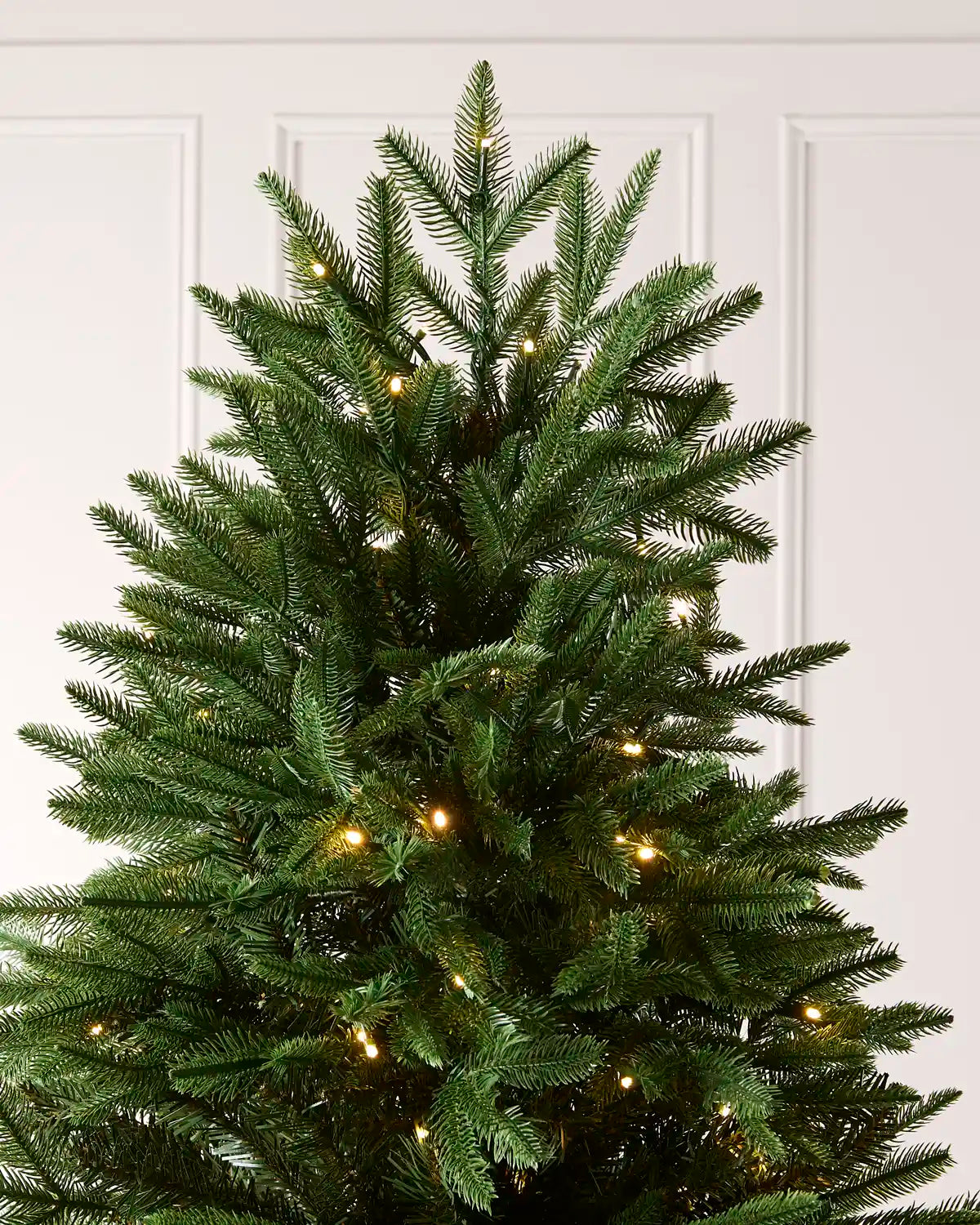 A green artificial Christmas tree with warm white string lights, shown against a white paneled wall background.