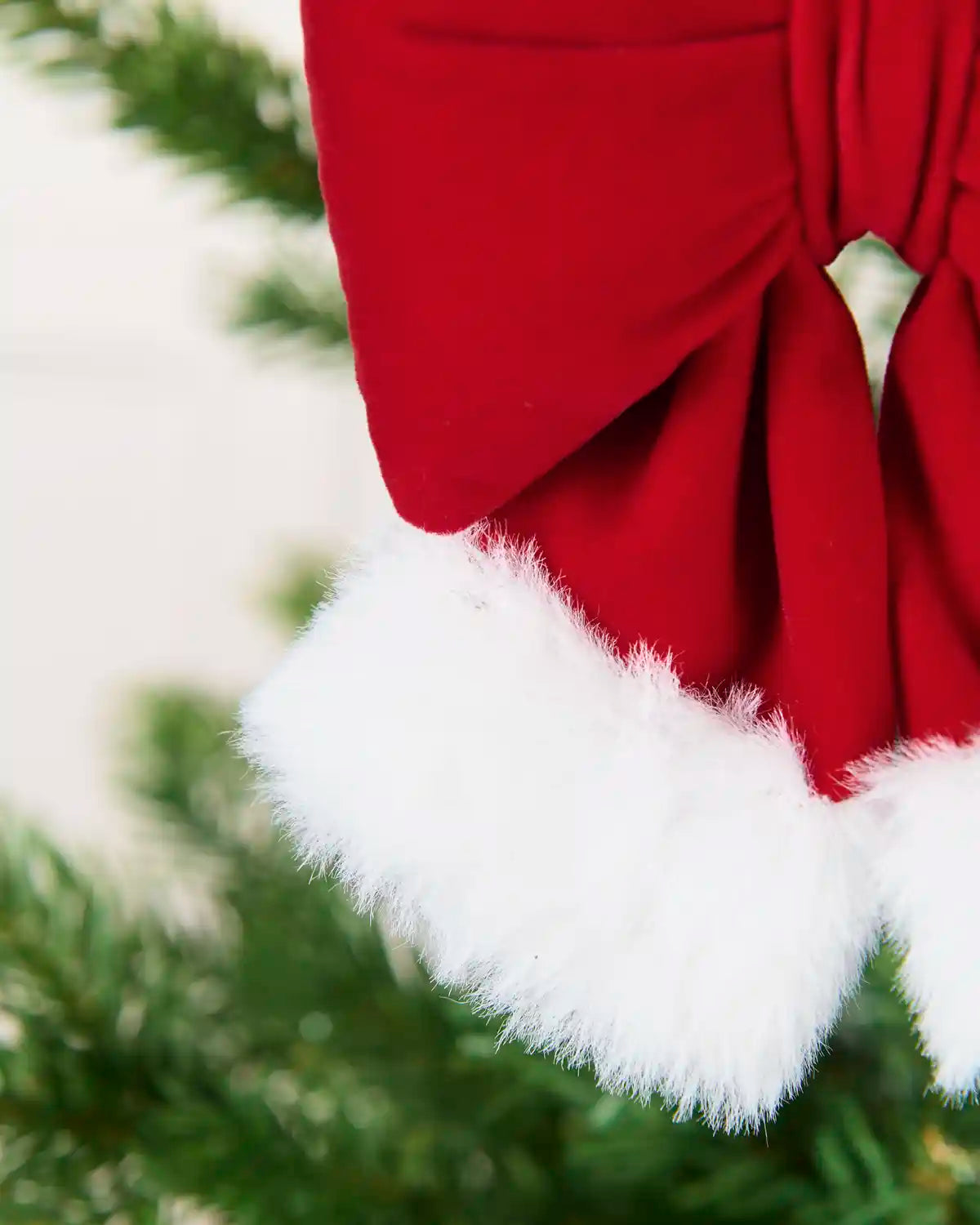 Close-up of Red and White Mini Velvet Santa Bow showing the white fluffy cuff with blurry Christmas tree branches in the background