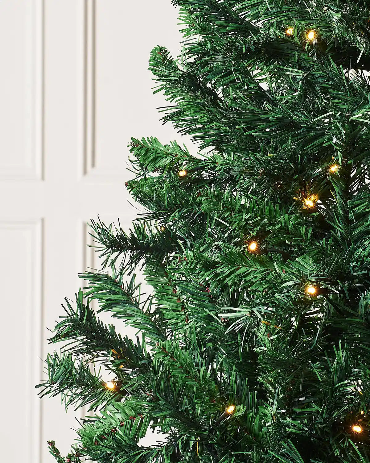 Close-up of a 7ft green artificial Christmas tree with lights against a cream panel wall.
