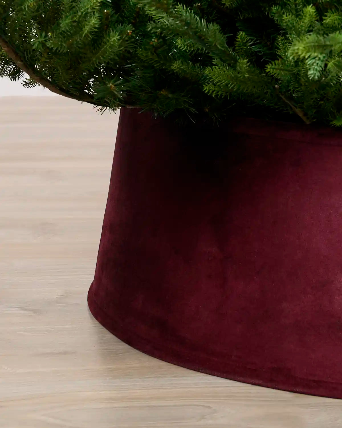 Close-up of a Burgundy velvet tree skirt holding a green artificial Christmas tree, set on a light wood floor.
