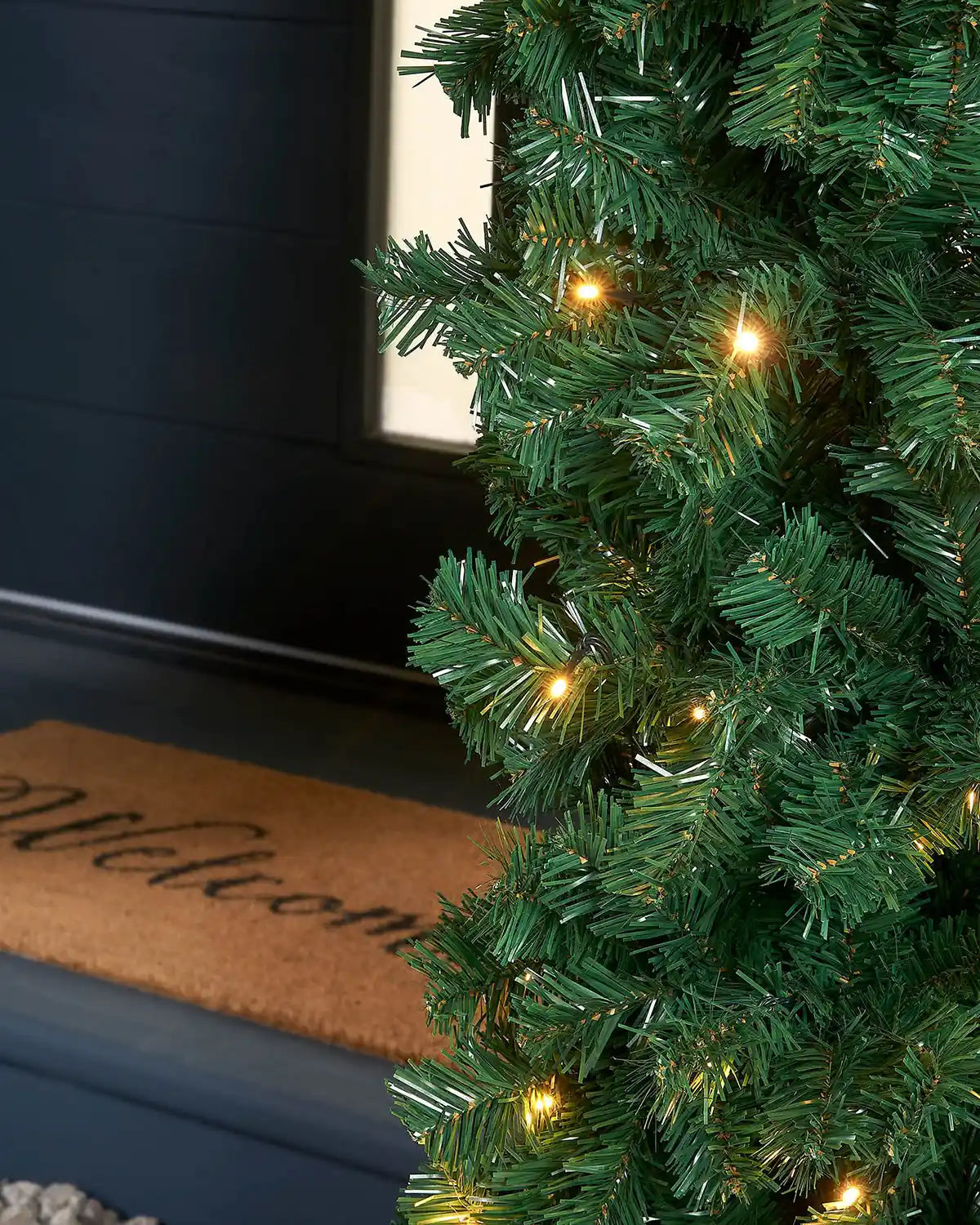 Close-up of a Christmas garland arch with lights against a black front door and a welcome door mat