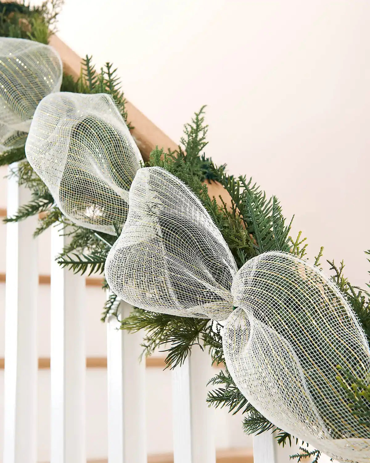 Close-up of a Christmas garland on a staircase banister with white and gold mesh ribbon against a neutral background
