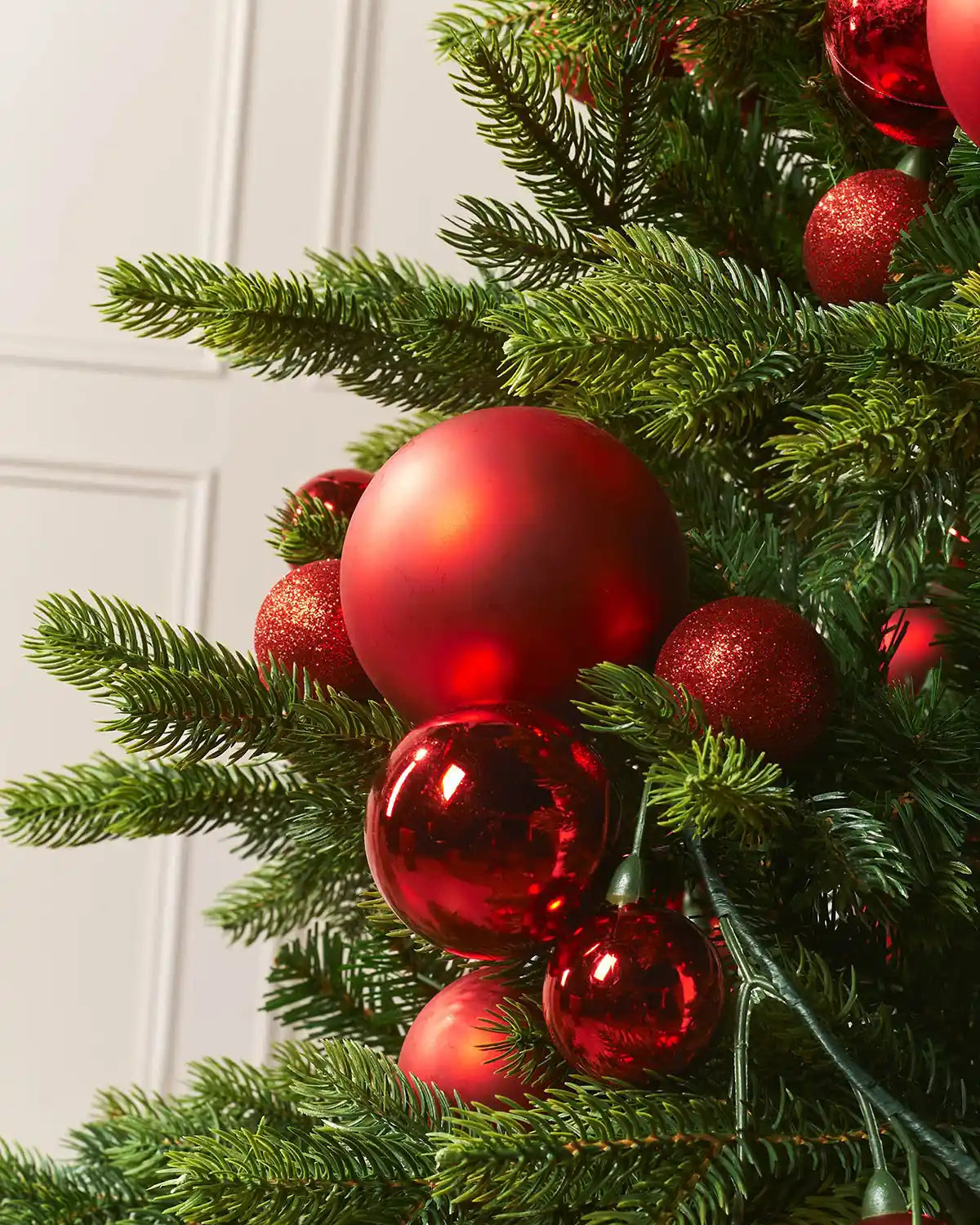 Close-up of a Christmas tree branch with red bauble garland against a white wall.