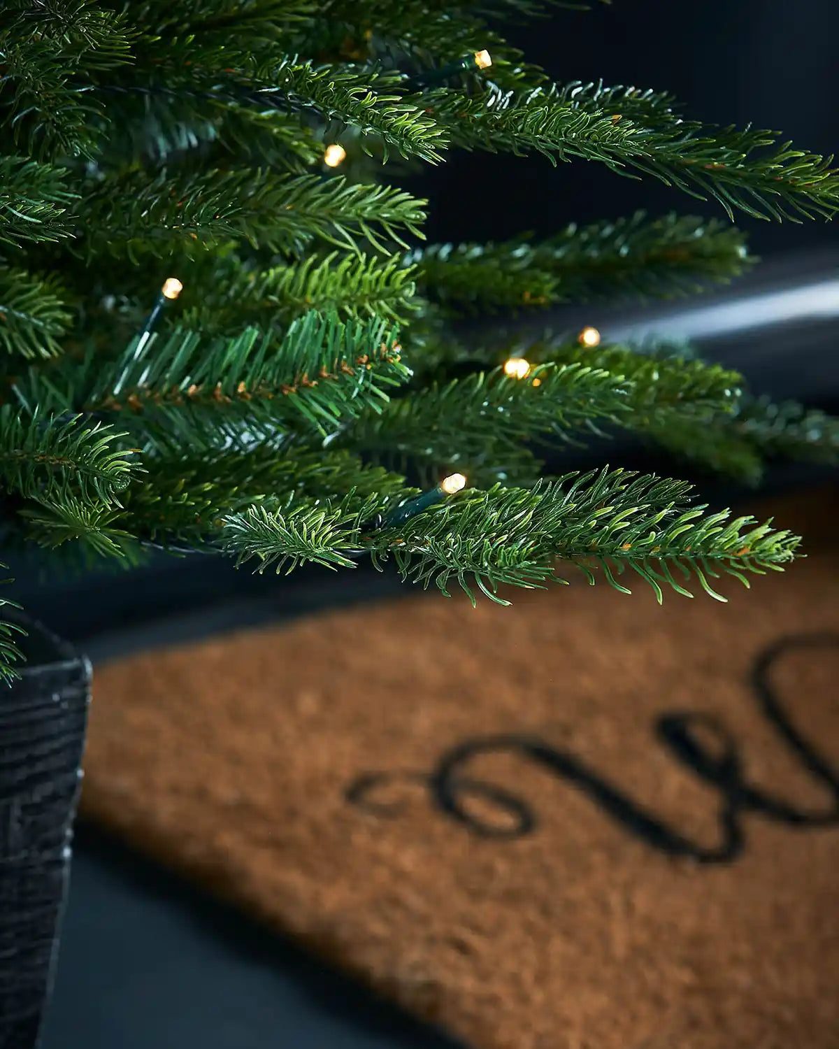 Close-up of a Decorated Pre-Lit Grand Fir Potted Christmas Tree with lights in a textured black pot on a doorstep with lights on a doormat.