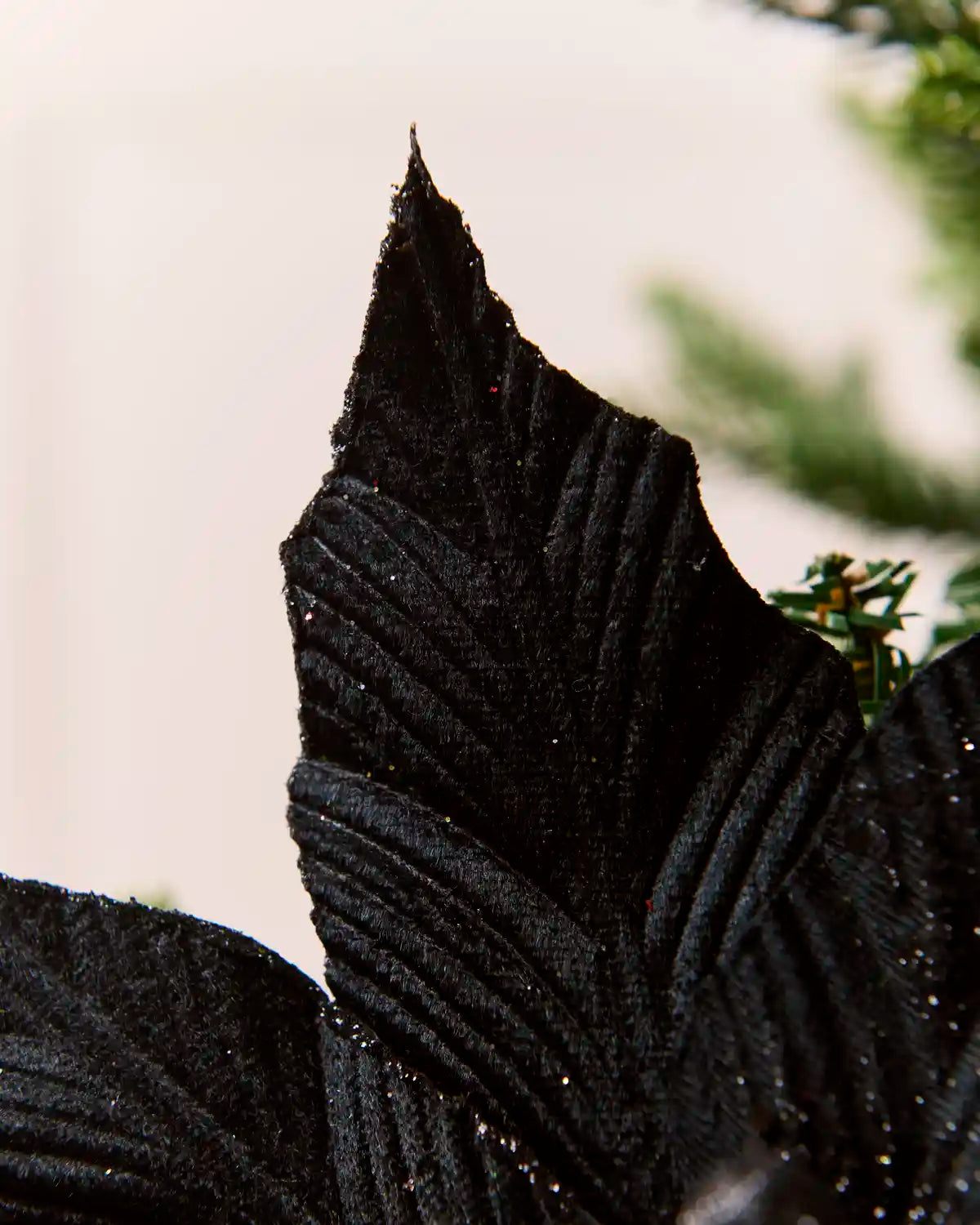 Close-up of a black textured artificial leaf with subtle glitter, shown against a soft off-white background with blurred green foliage.