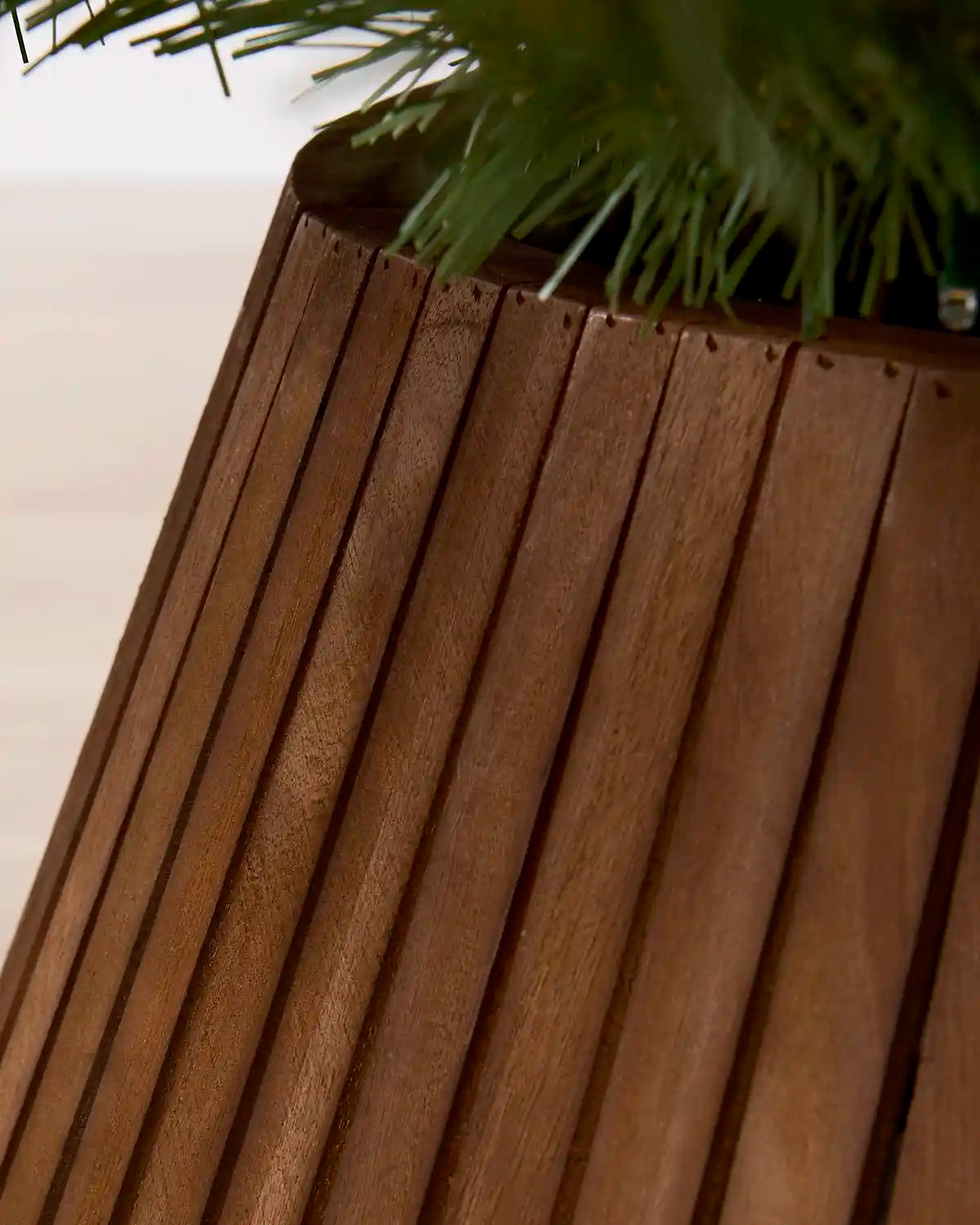 Close-up of a dark brown Christmas tree collar with vertical wooden slats in a medium brown finish, with green plant foliage visible at the top against a light neutral background.