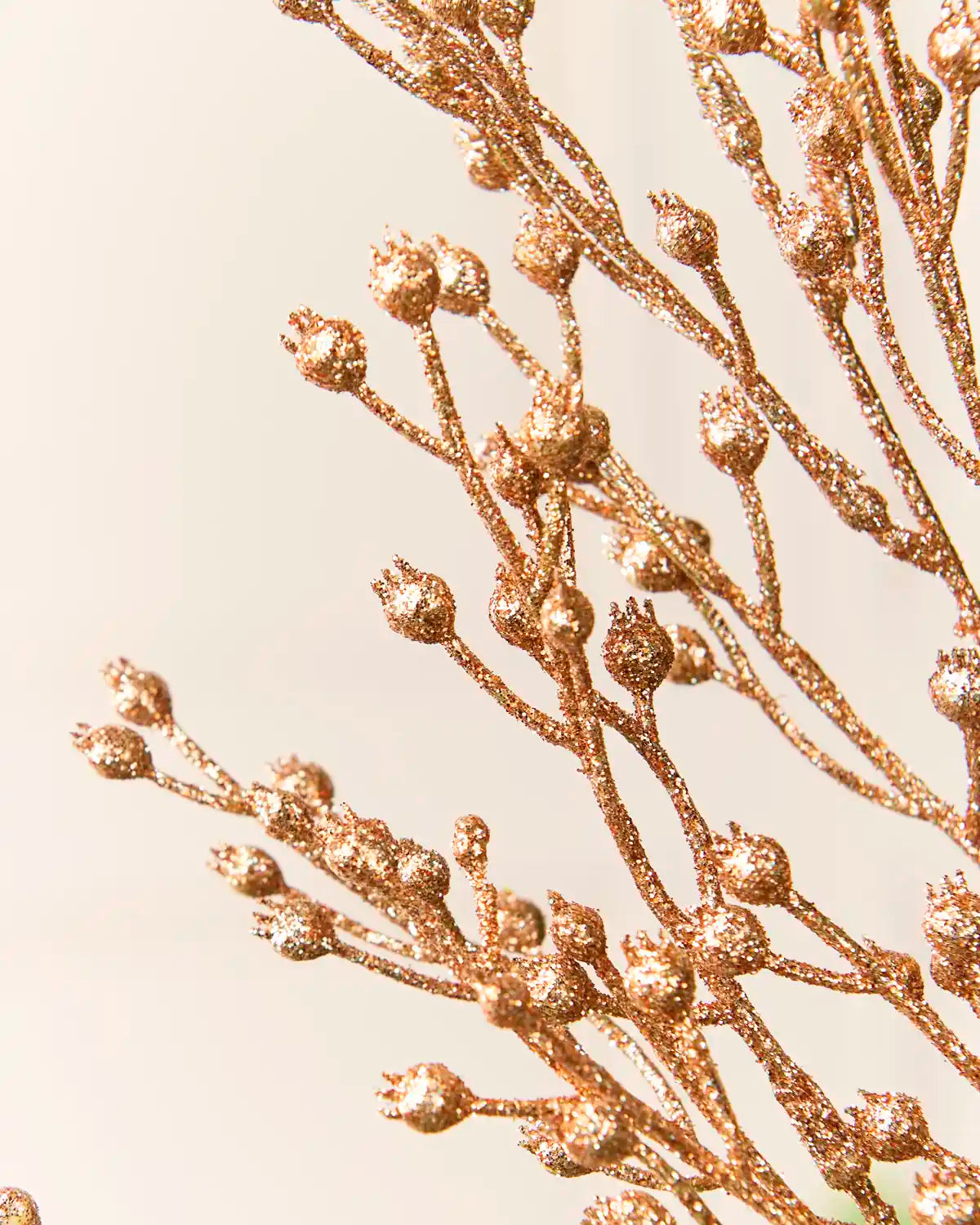 Close-up of a decorative branch arrangement covered in glittery rose-gold buds and stems against a soft cream background.