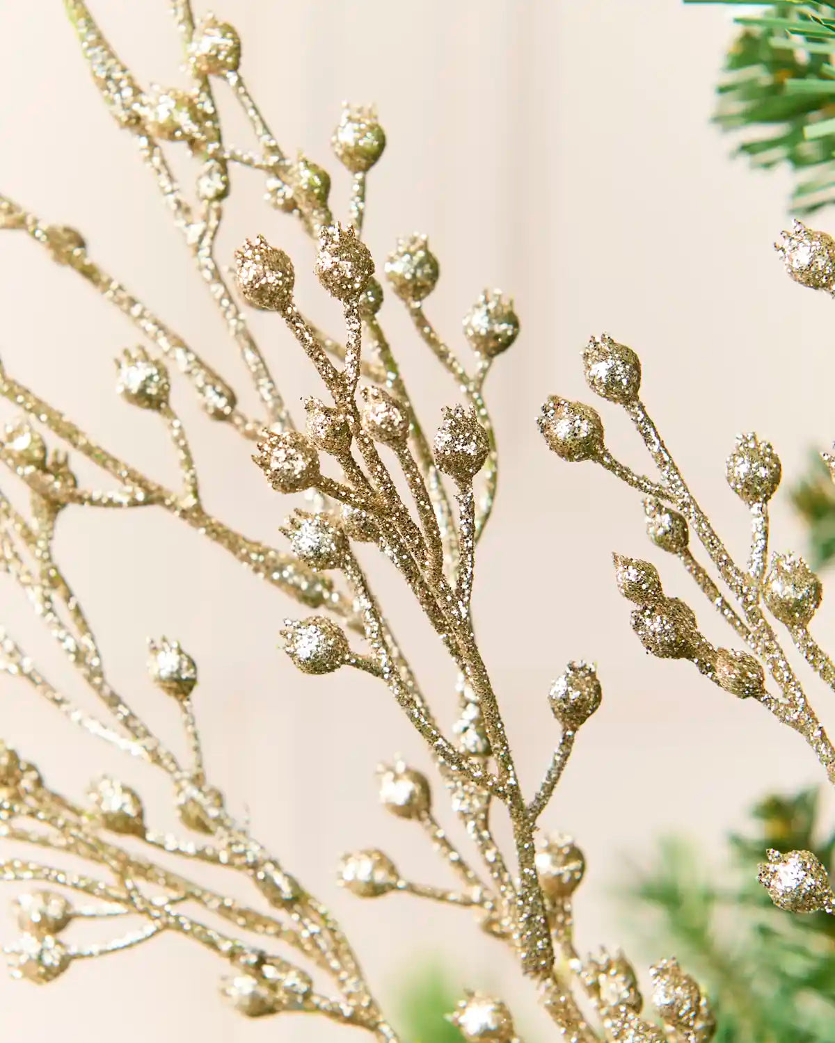 Close-up of a decorative gold glitter branch with small rounded buds, shown against a soft beige background with a hint of green foliage at the edges.