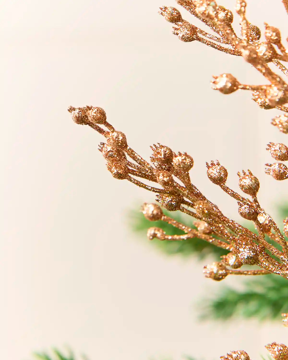 Close-up of a decorative rose gold glitter pine branch with small berry-like buds against a soft cream background with blurred green foliage.
