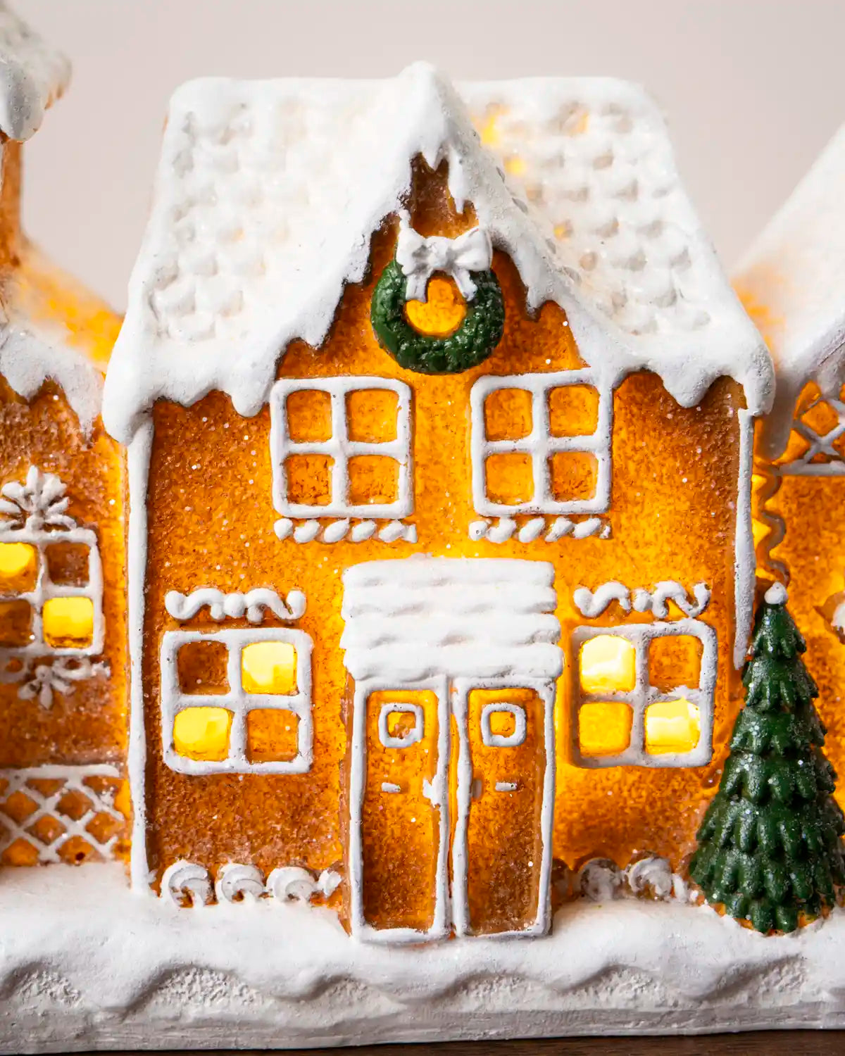 Close-up of a glowing gingerbread house with a snow-covered roof, white icing trim, illuminated windows, a green wreath, and a miniature Christmas tree on a snowy base