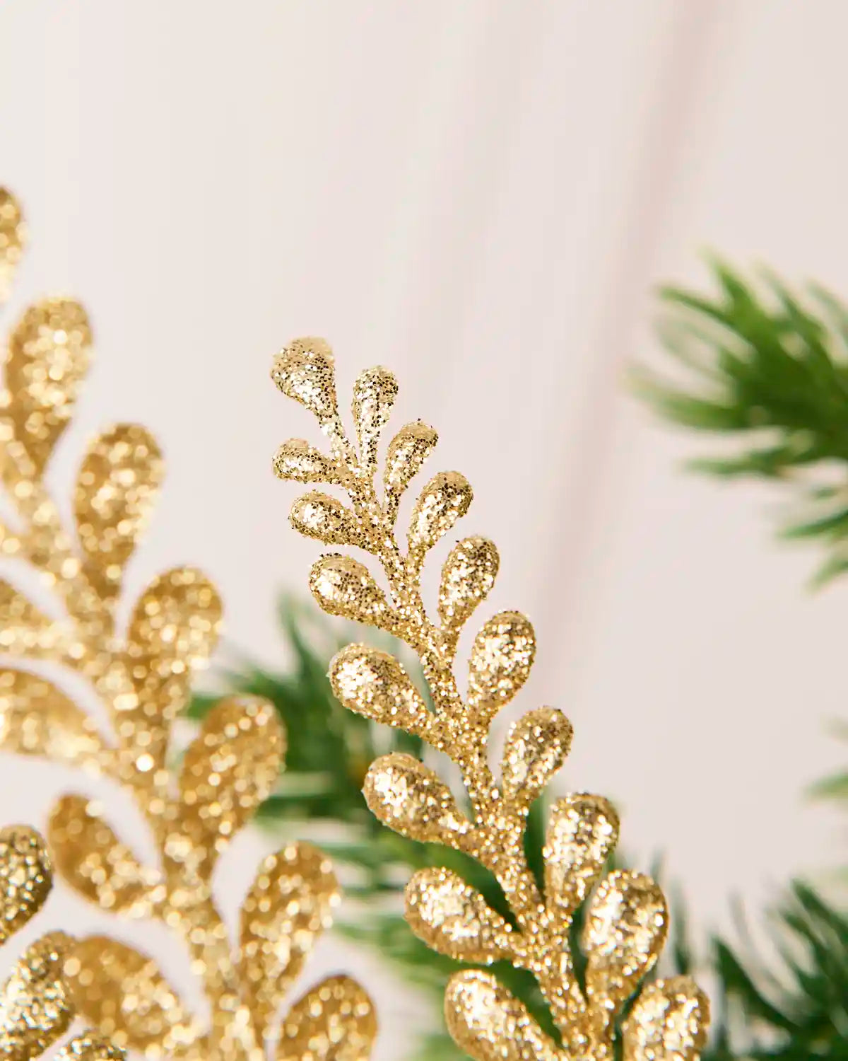 Close-up of a gold glitter leaf spray featuring a single detailed glitter-covered leafy stem with Christmas tree branches blurred in the background