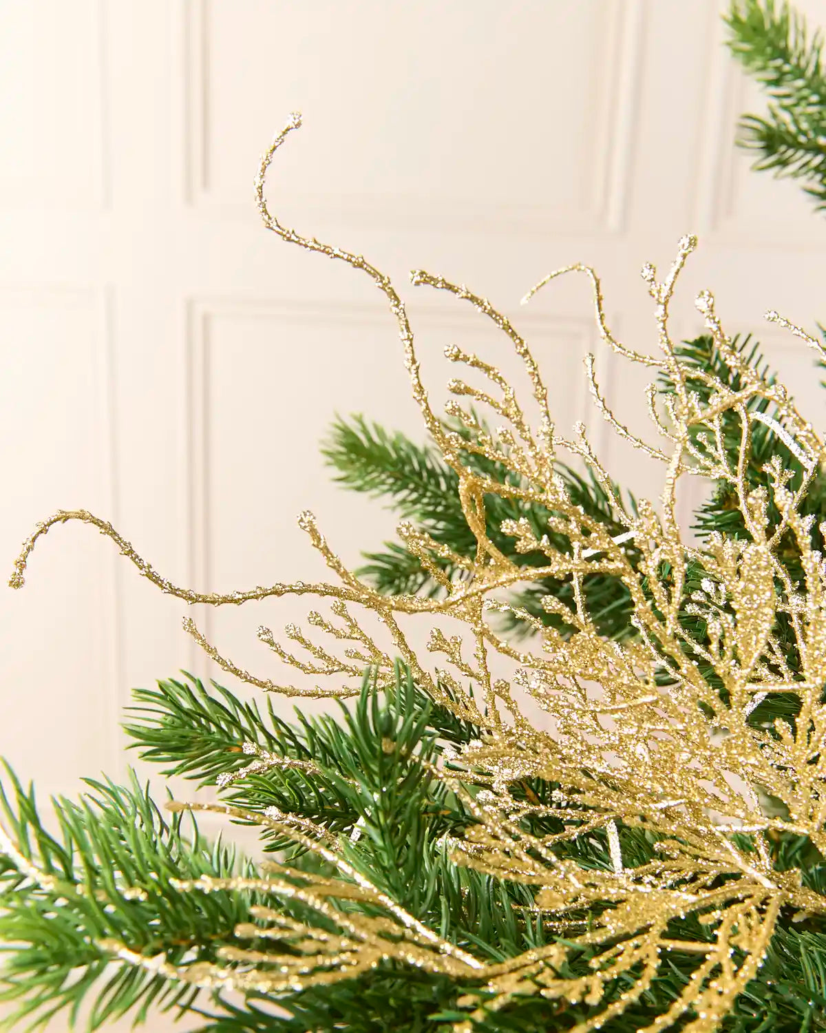 Close-up of a gold glittery decorative branch coral-like ornament layered over green pine branches against a soft cream background.