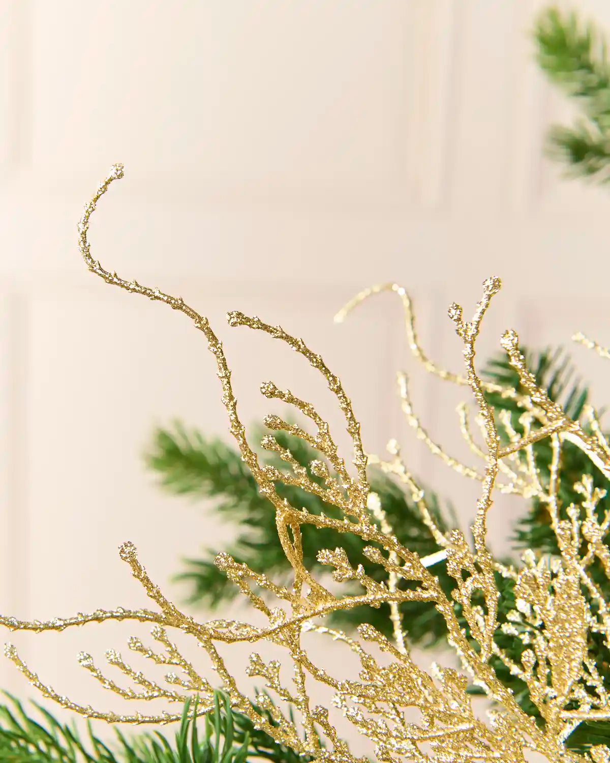 Close-up of a gold glittery decorative branch or artificial coral-like ornament layered over green pine branches against a soft cream background.