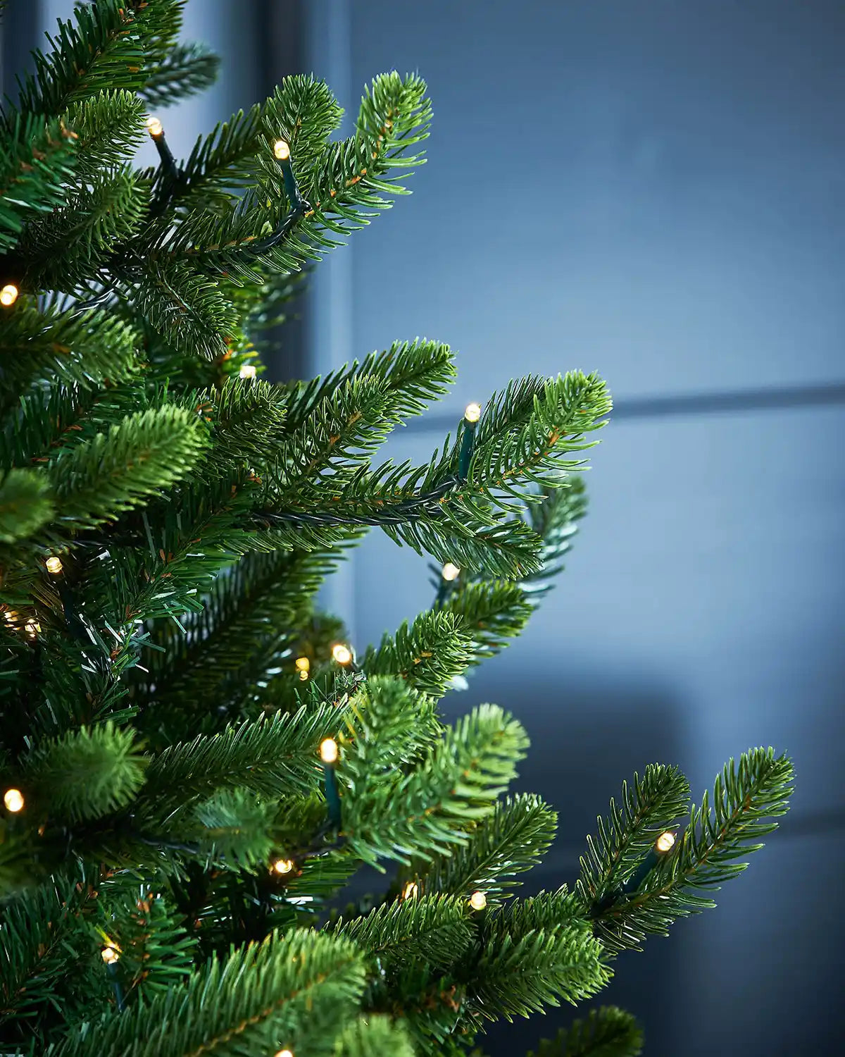 Close-up of a green artificial Christmas tree with single lit lights against a dark blue front door 