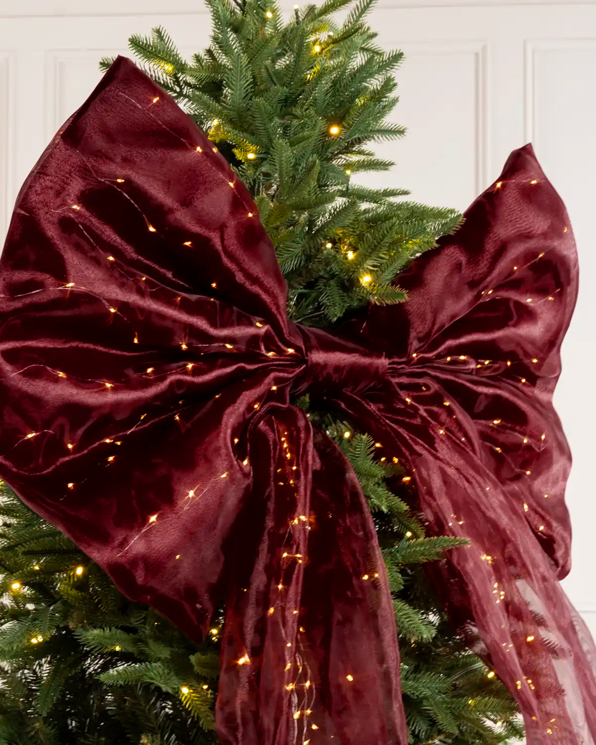 Close-up of a large burgundy Christmas bow with warm white lights, centered on a Christmas tree.