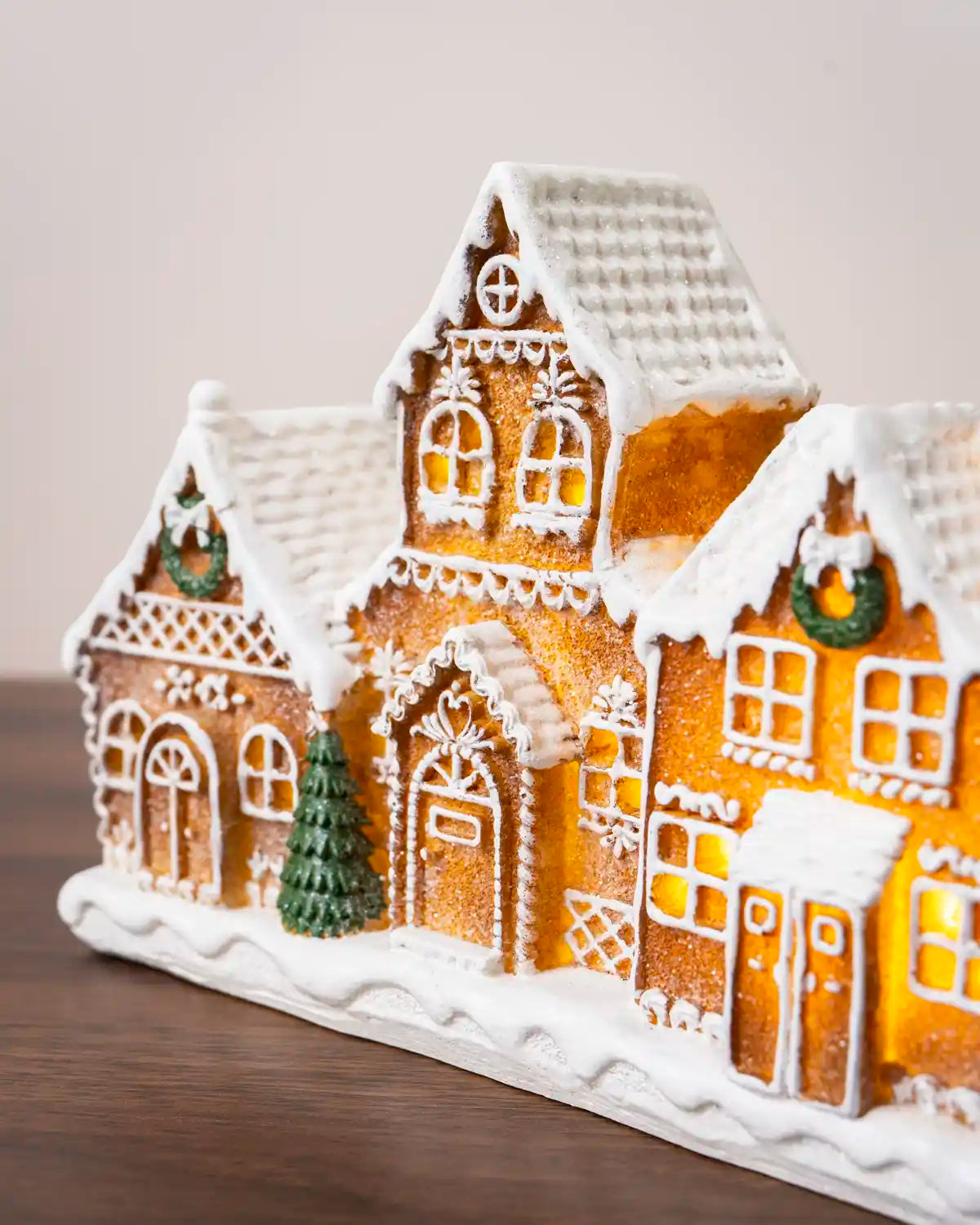 Close-up of a pre-lit gingerbread village showing detailed gingerbread houses with white icing, snow-covered roofs, glowing windows, green wreaths, and a miniature Christmas tree