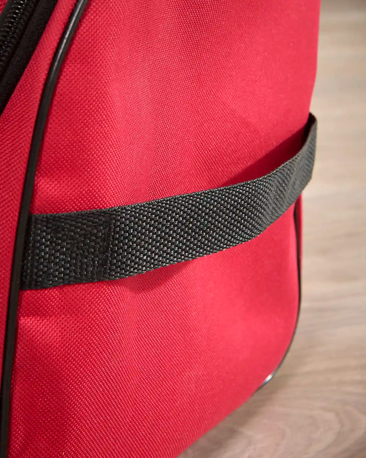 Close-up of a red fabric bag with a black woven strap across the front, photographed on a light wood surface.