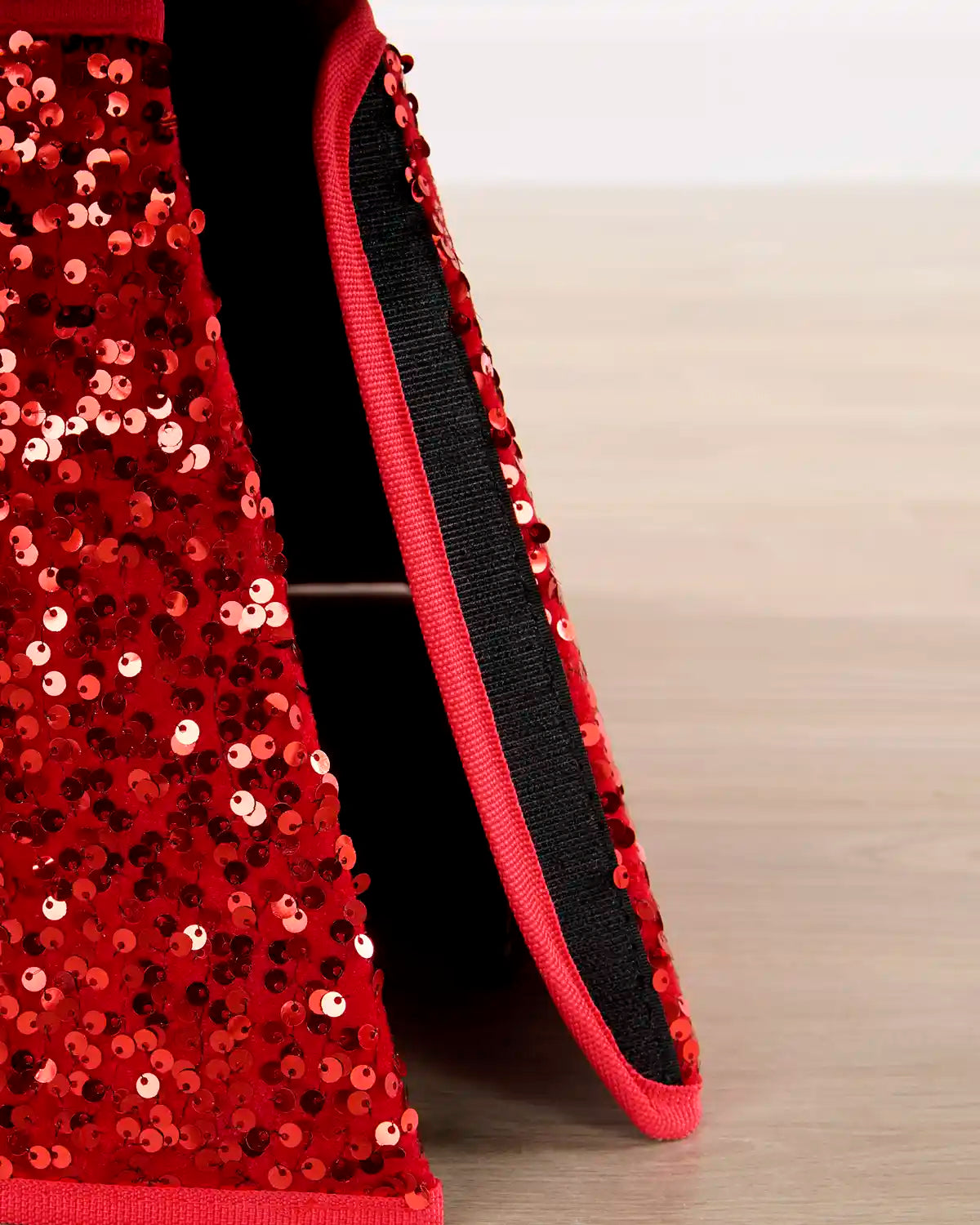 Close-up of a red sequined garment, likely a skirt or dress, showing the textured sequin surface, a red trim, and a black inner Velcro strip on a light wood floor.